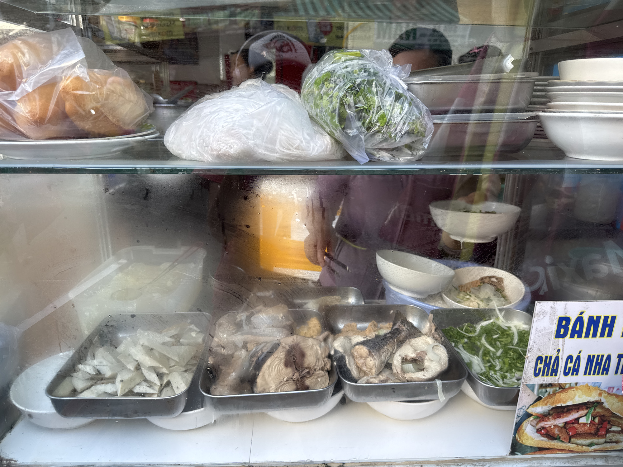 The image captures a street food stall in Nha Trang Ward, Vietnam, showcasing various food items displayed behind a glass enclosure. On the top shelf, several round bread rolls are visible, some encased in plastic bags. Next to them, a large plastic bag contains what appears to be shredded white noodles or rice paper, and another bag holds a bundle of green leafy vegetables, possibly herbs. A stack of white bowls and plates are arranged on the right side of the top shelf.
The middle shelf reveals more food items in metal trays. One tray contains sliced fish cooked in broth, while another holds more pieces of cooked fish. A bowl filled with a light-colored liquid is also visible.
On the bottom shelf, a metal tray holds what looks like thinly sliced white root vegetables, possibly taro or radish, submerged in liquid. To the right, a metal container is filled with chopped green onions and possibly other herbs. A sign with Vietnamese text and an image of a sandwich is placed in the foreground on the right. The overall impression is that of a well-stocked stall preparing for business, with a focus on fresh ingredients and seafood. The lighting suggests it is daytime, and the reflections on the glass indicate an outdoor setting.