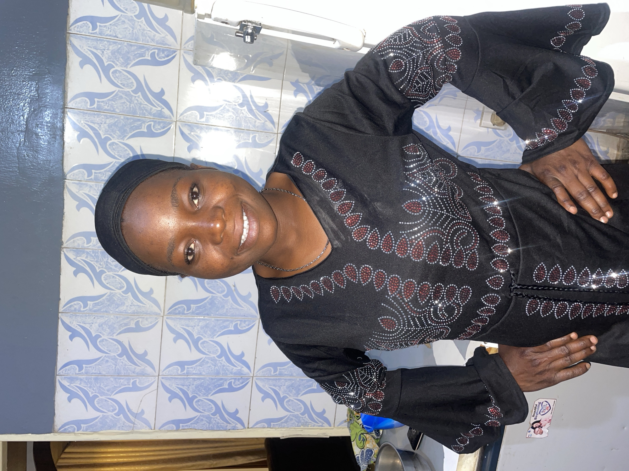A woman with dark skin and short hair is smiling at the camera. She is wearing a black dress with decorative embellishments. She is also wearing a black head covering. The background shows a wall with blue and white tiles. There's a sink and some other objects on a counter. Based on visual clues and the prompt, the image is assumed to be taken in Jos, Nigeria.