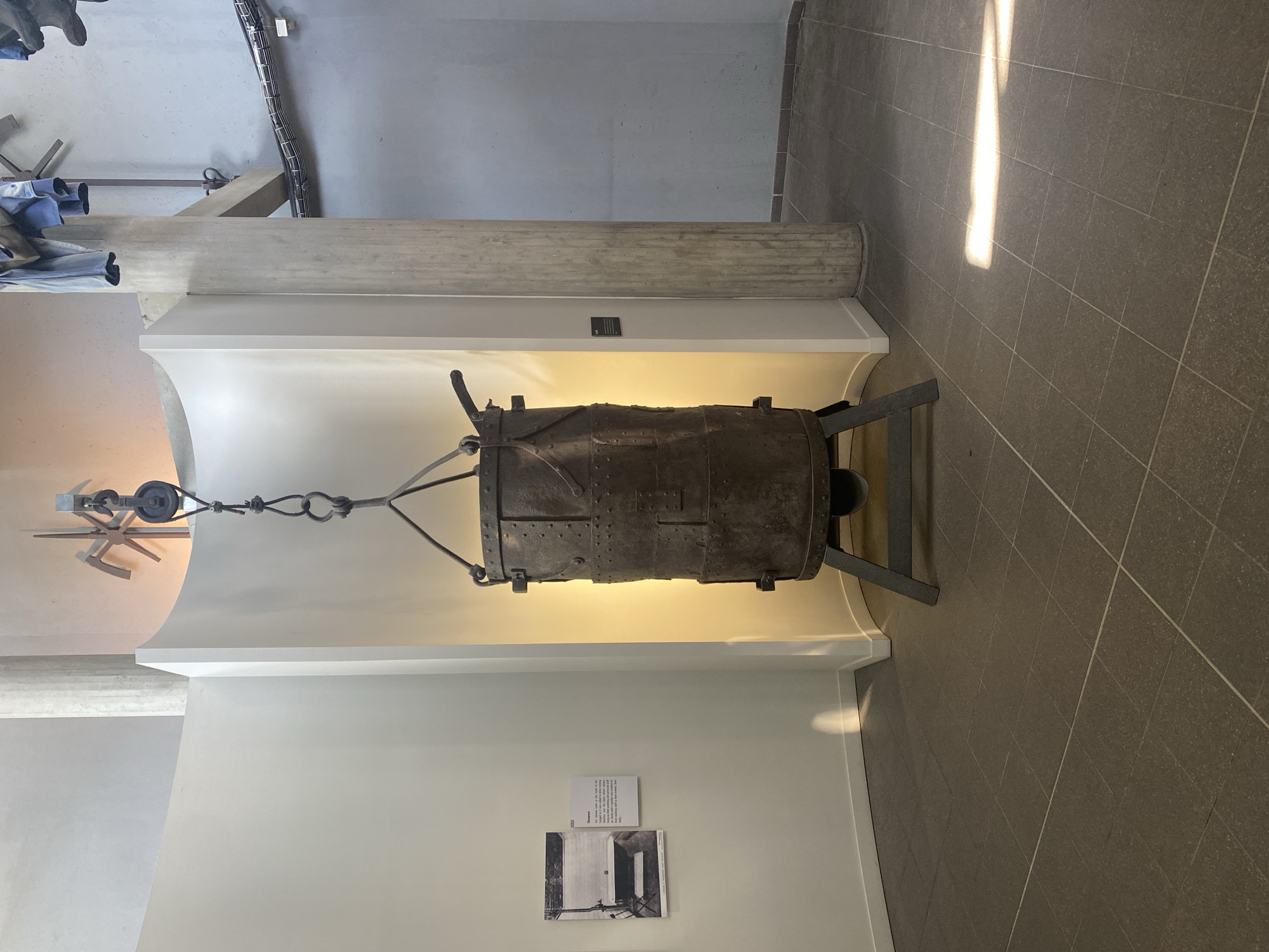 Indoor shot featuring an old mining artifact in La Motte-d'Aveillans, France. A cylindrical metal object, rusty and aged, is suspended from a pulley system attached to a white architectural structure. The device has visible bolts and a support stand on the right. It is displayed against a curved white wall. A framed description and historical photo are on the wall below the artifact. The floor is tiled in a dark gray tone, reflecting light. The overall scene is a museum or exhibition area.
