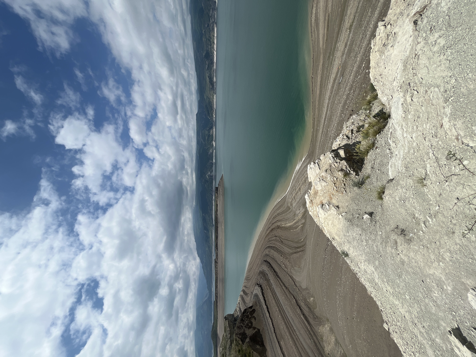 The image shows the landscape of Chirkei, Russia. The view is of a large body of water, a reservoir possibly formed by the Chirkei Dam. The water is turquoise-green in color. The banks of the reservoir are visible, revealing extensive soil and rock formations marked with striations, indicating varying water levels over time. To the left, mountains or hills rise in the distance, partially obscured by clouds. The sky is blue with scattered white clouds. A rocky cliff edge forms the foreground on the right side of the image, with sparse vegetation. The photo is taken from an elevated position.