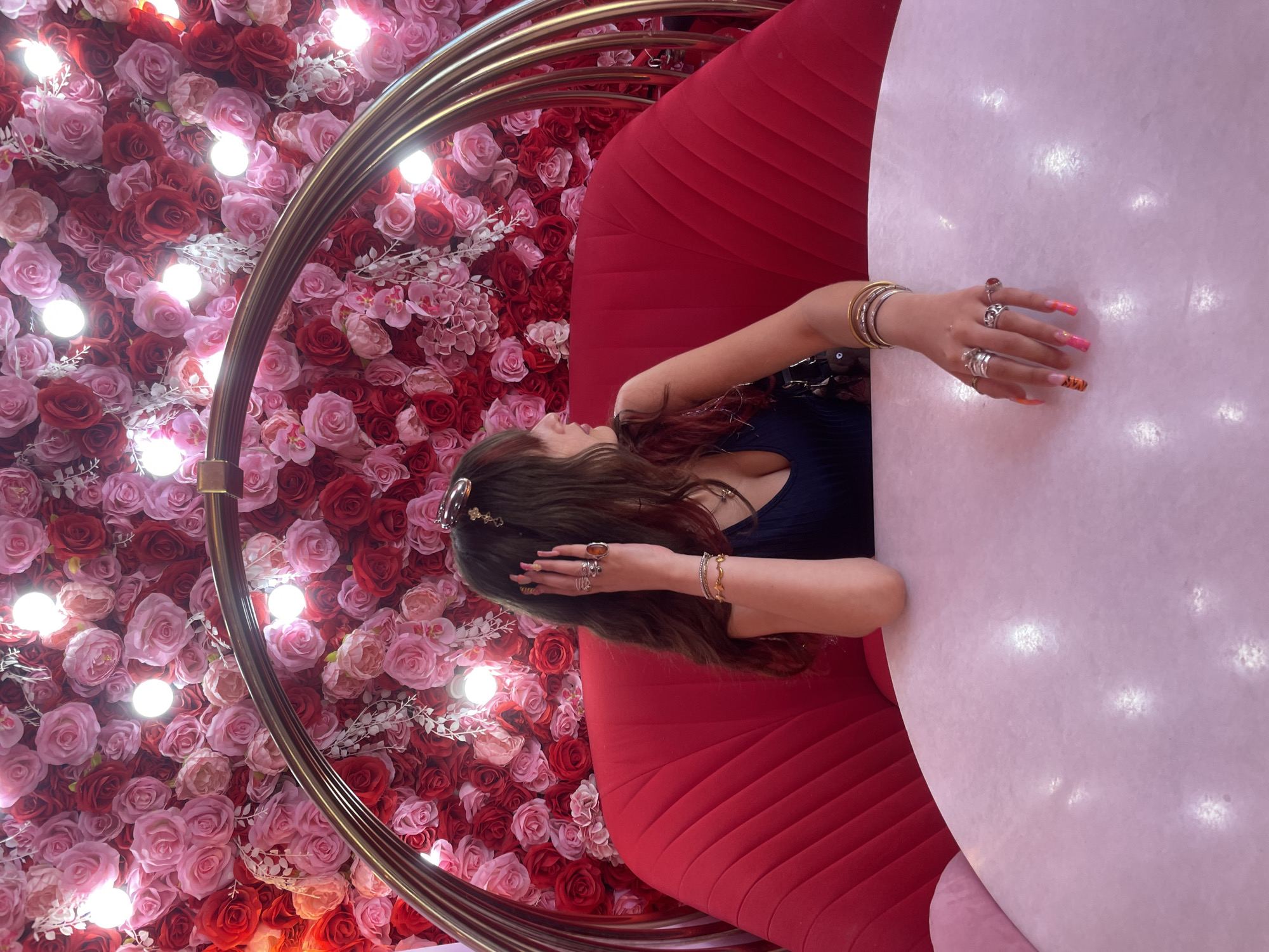 In Paphos Municipality, Cyprus, a young woman with long, dark hair and bangs sits at a table in front of a wall of red and pink roses. She is wearing a navy blue top with a low neckline and several rings on her fingers, including one with a large orange stone. She also has several bracelets on her wrist and long, brightly colored nails. The woman is looking to her right, with her hand resting on her head. She is seated on a plush red chair that is part of a circular, gold-colored hanging seat. The table in front of her is round and pale pink, with a marble-like surface. The background is a dense arrangement of artificial roses, interspersed with small, warm-toned lights.