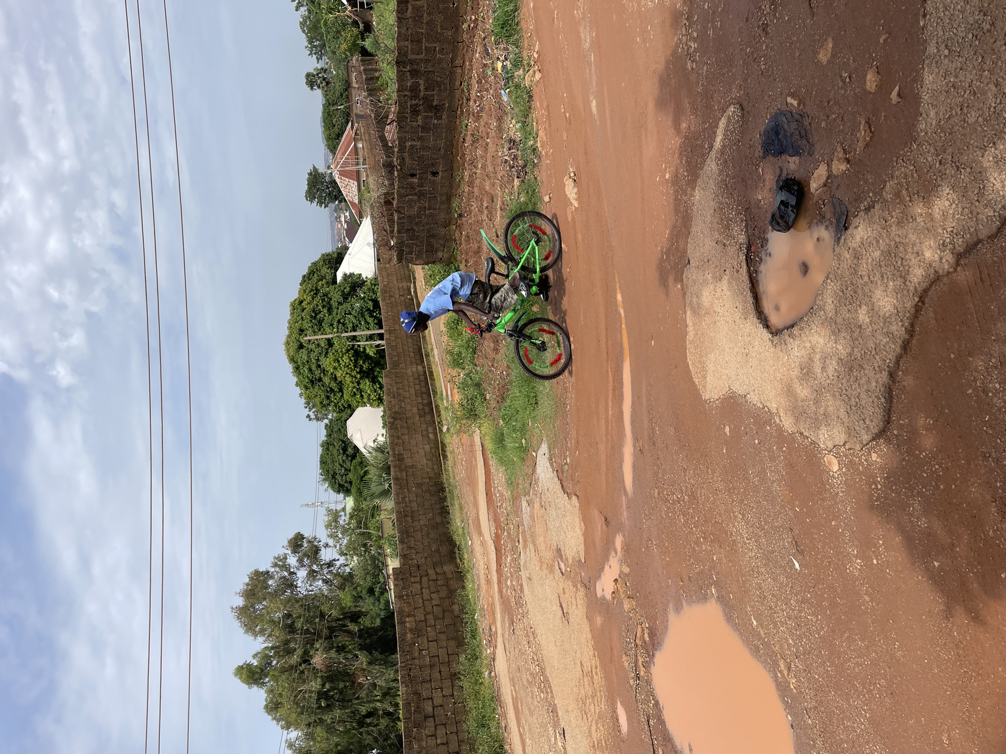 A young boy wearing a blue t-shirt and blue cap rides a bright green bicycle with red accents on a muddy, unpaved road in Jos, Nigeria. The road is riddled with large potholes filled with murky water, and the surrounding ground is a mix of red dirt and gravel.
In the background, a brick wall runs alongside the road, separating it from a grassy area with trees. Beyond the wall, several houses with distinctive orange, pitched roofs are visible, interspersed with lush green trees. A white tent-like structure is also present among the buildings.
The sky is bright and partly cloudy, suggesting daytime. The overall scene depicts a rural or semi-urban environment, characterized by unpaved infrastructure and natural elements. The boy appears focused on his ride, navigating the challenging terrain. There is no visible text in the image.