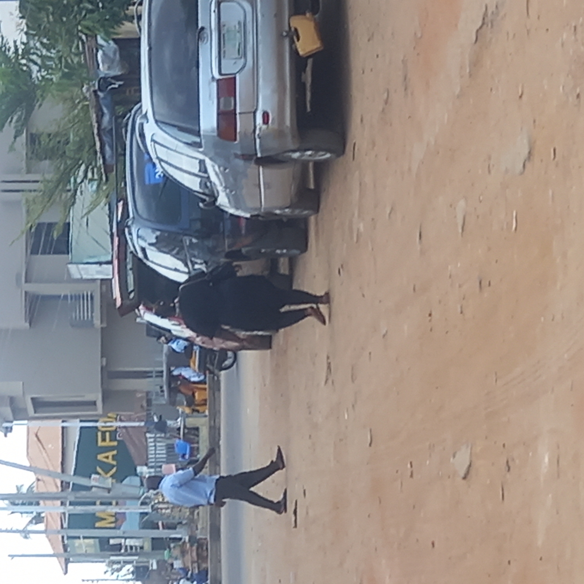 The image depicts a street scene in Dadi Riba, Nigeria. Several cars are parked along the side of a dirt road. A woman in dark clothing walks past the cars. A man in a light blue shirt and dark pants stands on the road with one arm raised. A building with a sign reading "MKAFO" is visible in the background. Other individuals and motorcycles are present in the scene. The ground is mostly a dirt road.
