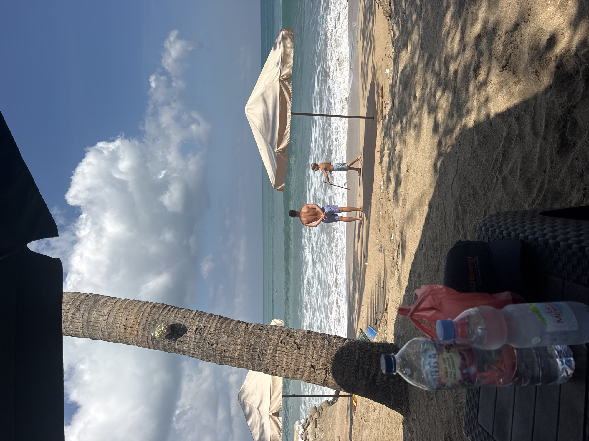 The image depicts a tropical beach scene in Kuta, Indonesia, under a bright blue sky with scattered white clouds. In the foreground, a table displays two water bottles and a red plastic bag. A large palm tree trunk dominates the left side of the frame, its rough bark texture visible. Beyond the tree, the sandy beach stretches towards the turquoise ocean. Two beach umbrellas provide shade on the sand, one to the left and a larger, tan one in the middle ground. Two individuals are visible on the beach. A man, seen from the back, stands facing the ocean, wearing blue patterned swim trunks. To his right, a young boy, also facing the ocean, holds a stick. The waves are breaking gently on the shore, creating white foam. The overall impression is a relaxed day at the beach, with elements of everyday life on display.