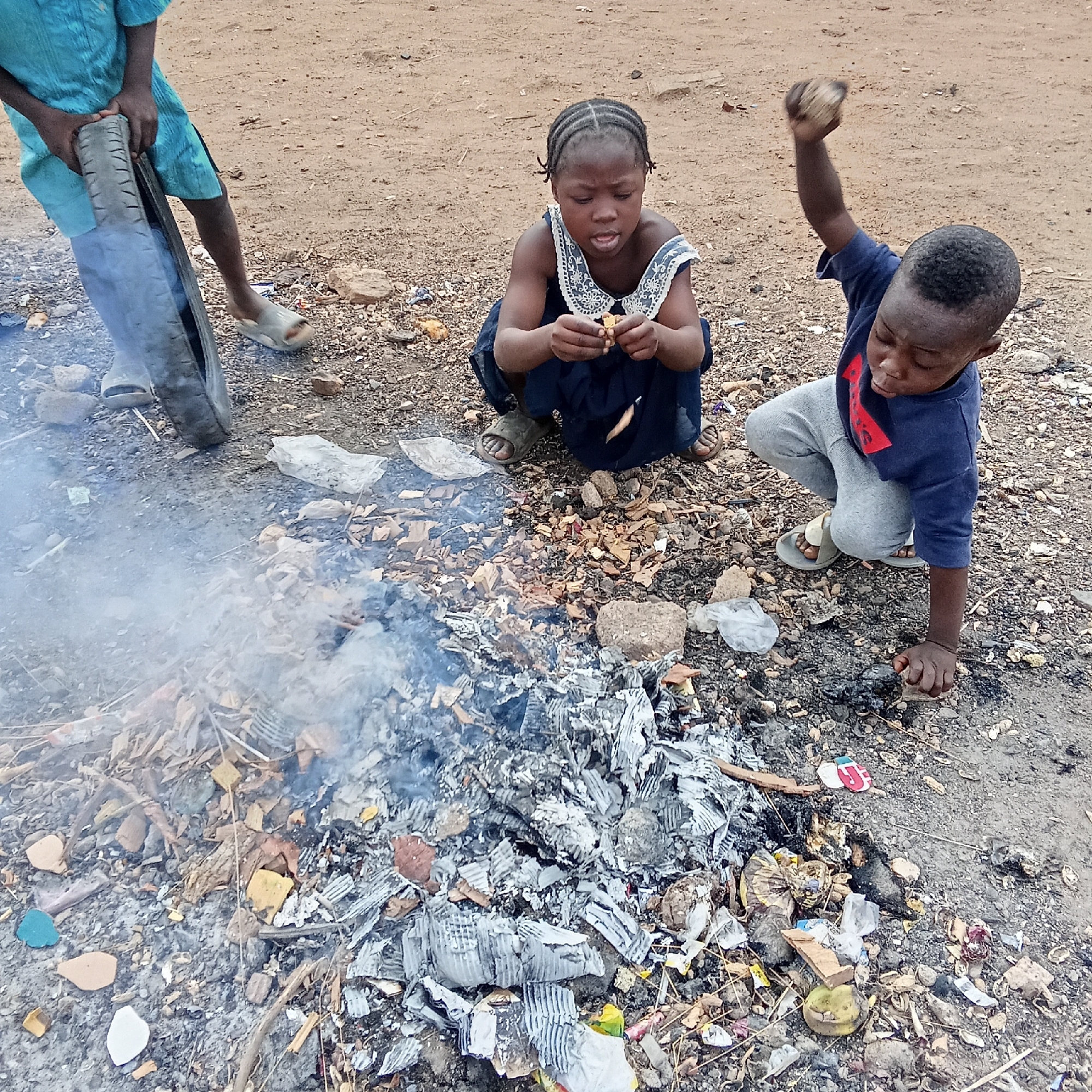 Three children are gathered around a fire in Akwanga, Nigeria. One child stands to the left, holding a tire. Another child is crouched down, picking at something. The third child is on his knees, with his right hand on the ground and his left arm raised, holding a rock. A fire burns in front of them, surrounded by ash, debris, and litter scattered on the ground. The backdrop is a dirt ground.