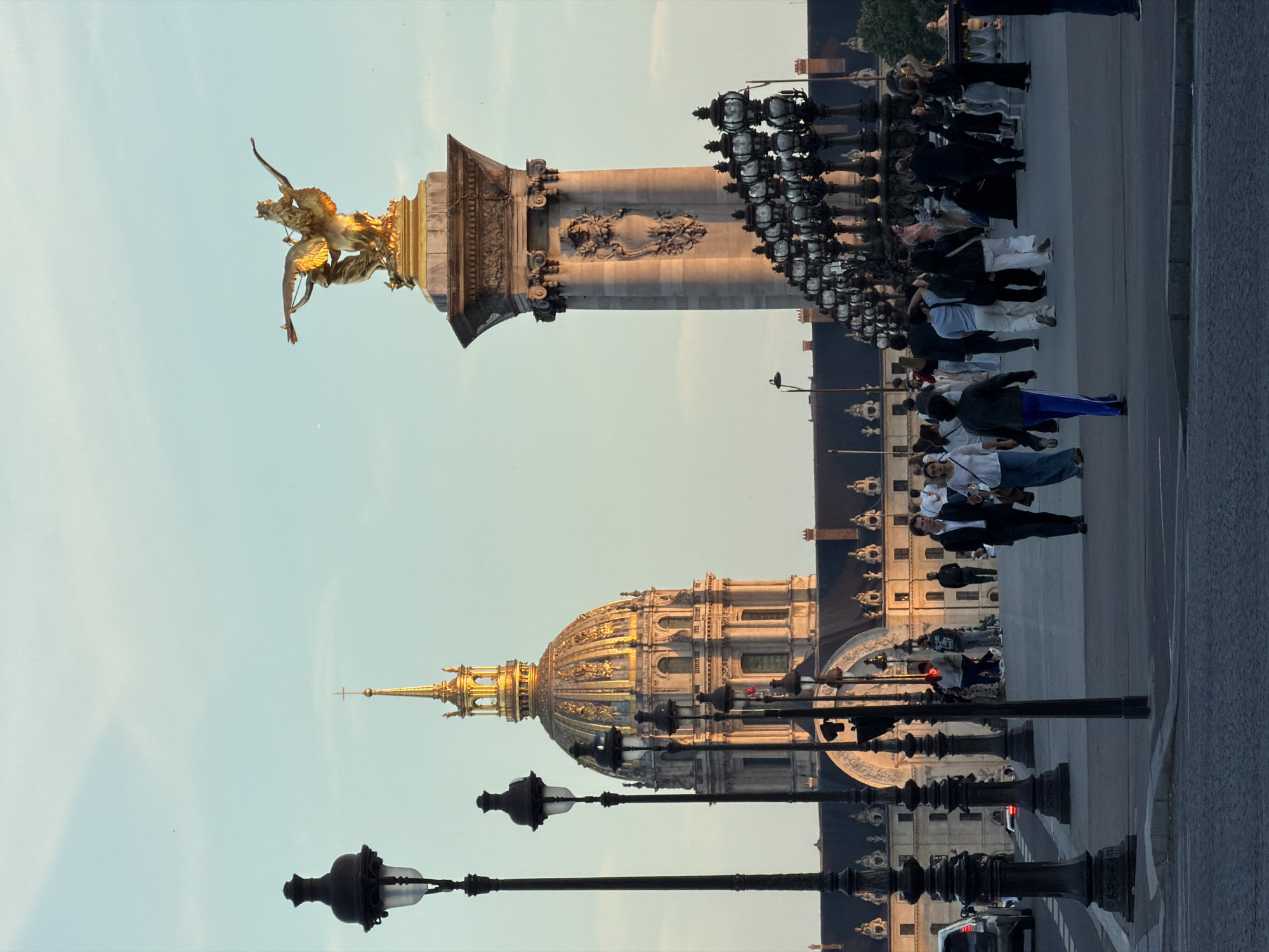 The image captures the Alexandre III Bridge in Paris, France. The bridge is depicted with its ornate lamp posts lining the walkway. In the background is the dome of Les Invalides. People are walking along the bridge. The image also showcases one of the gilded sculptures that adorn the bridge's columns.