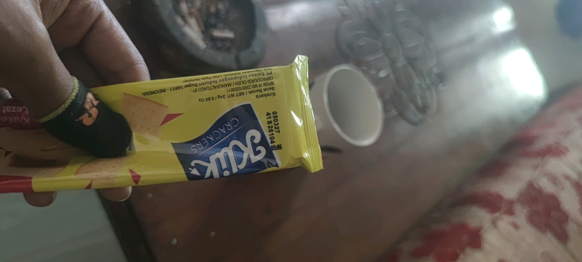 In Gambarsari, Indonesia, a person's finger, partially covered by a black finger cot with an orange graphic, is holding a yellow package of KREKERS crackers. The packaging displays the brand name "Kulik" in blue cursive font, with the word "CRACKERS" written above it. Text on the package indicates "MANUFACTURED BY: PT. Serena Indopangan Industri Bogor 16911 - INDONESIA," and "Berat Bersih / NET WT: 24g / 0.85 Oz." A batch number "050327" and a date "41S26104" are visible. The crackers are light brown and square-shaped. The background shows a blurry brown surface, possibly a table, with out-of-focus objects including what appears to be a mortar and pestle and a white cylindrical container. The overall lighting is dim, with some bright reflections suggesting artificial light sources.