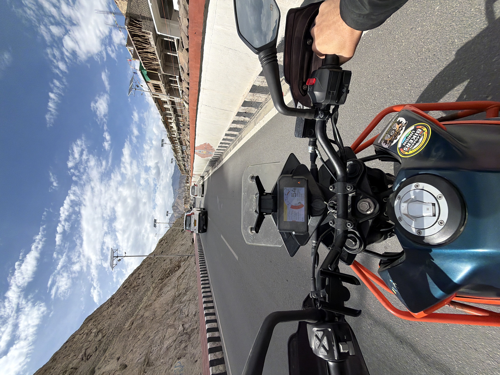 The image depicts a first-person perspective from a motorcycle on a paved road in the city of Leh, India. The motorcycle is a blue and orange KTM adventure bike. The road is bordered by a steep, rocky mountainside on the left and a construction site with unfinished buildings on the right. Several white vehicles are visible on the road ahead, including a white SUV and a white hatchback. The sky is blue with scattered white clouds. The motorcycle has a digital display showing a speed of 54. The rider's hand is visible on the right handlebar, wearing a dark jacket. The motorcycle has several stickers on its fuel tank, one of which reads "LADAKH BIKERS UNION". The scene appears to be during daylight hours.