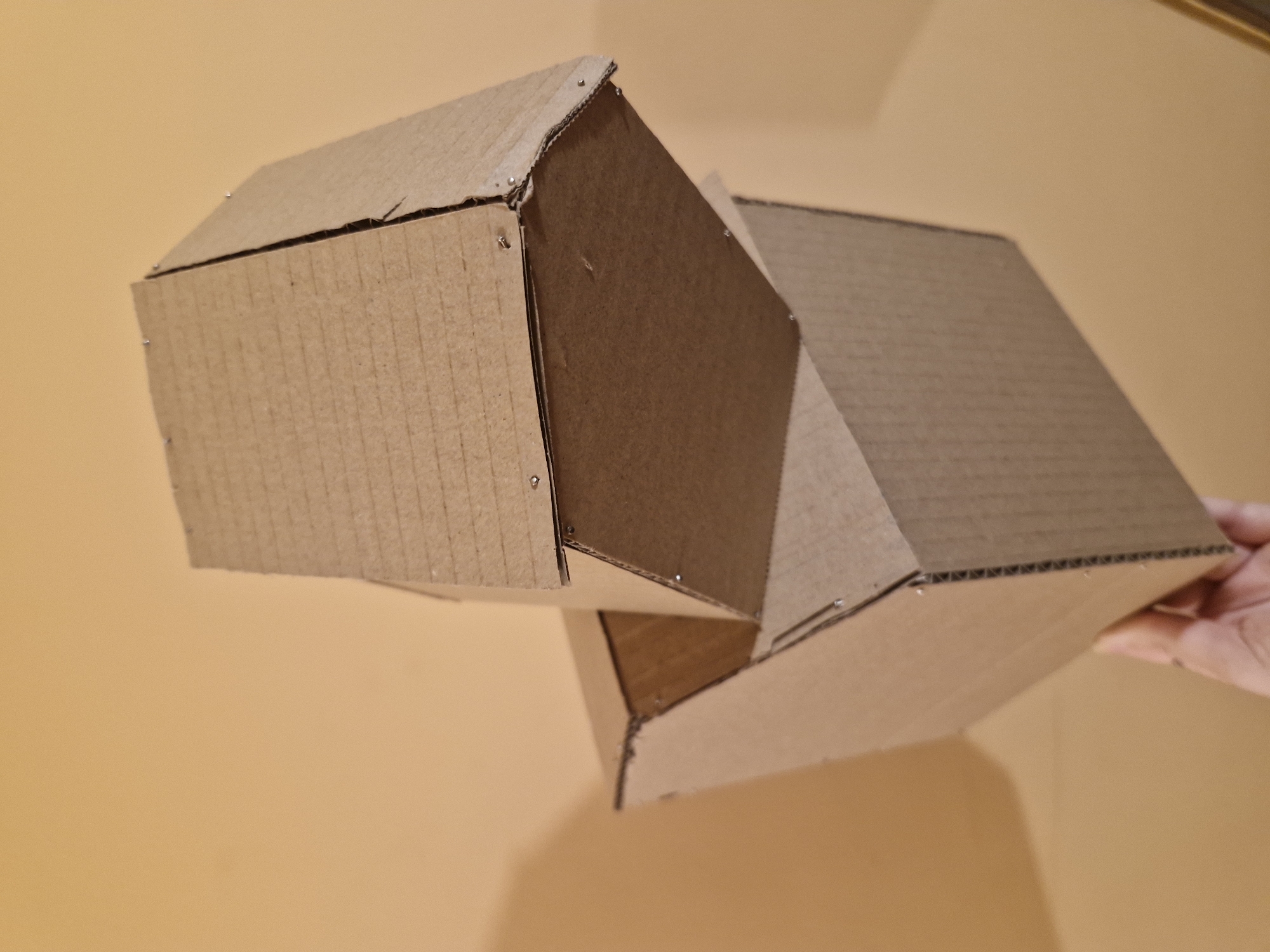 The image shows a handmade cardboard structure composed of multiple boxes connected at angles. The boxes appear to be held together with staples. The background is a plain, light orange surface, possibly a wall or tabletop. A hand is visible holding one of the boxes. The scene does not contain any information regarding Sofia, Bulgaria.