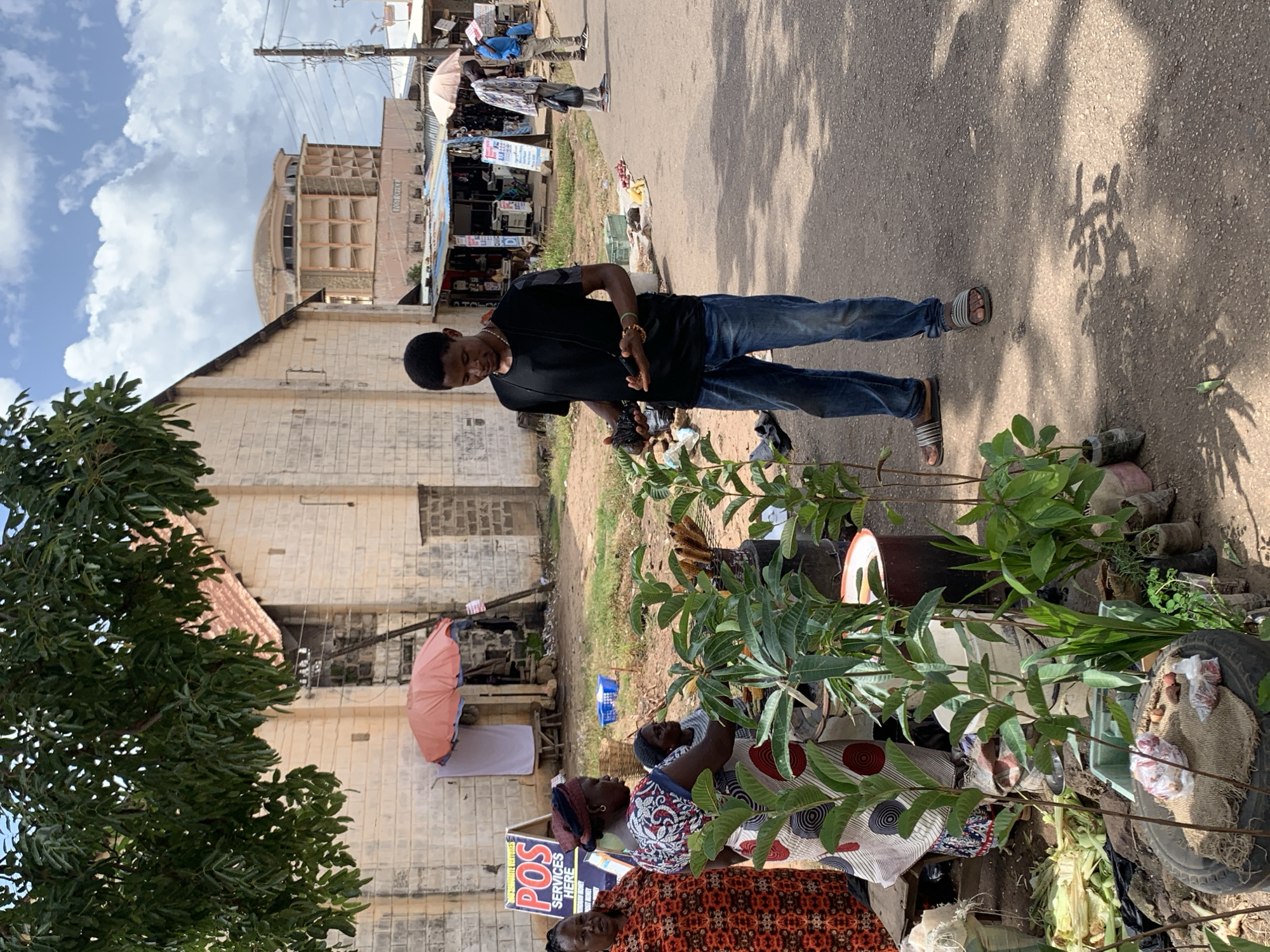 In Jos, Nigeria, an outdoor market scene unfolds on a sun-drenched street under a partly cloudy sky. A young man in a black t-shirt and blue jeans stands in the foreground, holding a black electronic device, possibly a camera, in his right hand. He is wearing sandals and has a lanyard around his neck. To his left, three women are gathered around a stall. One woman, wearing a vibrant orange and red patterned dress, stands on the far left. Next to her, a woman in a colorful patterned dress and headscarf is tending to what appears to be a plant stall. A third woman, partially visible, is also present.
The stall features numerous potted plants, some with dark soil and others in clear plastic bags. Various items, including what looks like corn husks, are arranged on the ground. In the background, several market stalls with corrugated metal roofs line the street, some displaying umbrellas and merchandise. A building with a light-colored brick facade and a red-tiled roof stands behind the stall, and further in the distance, a distinctive, rounded modern building with balconies is visible. The street is paved and appears to be a busy commercial area.