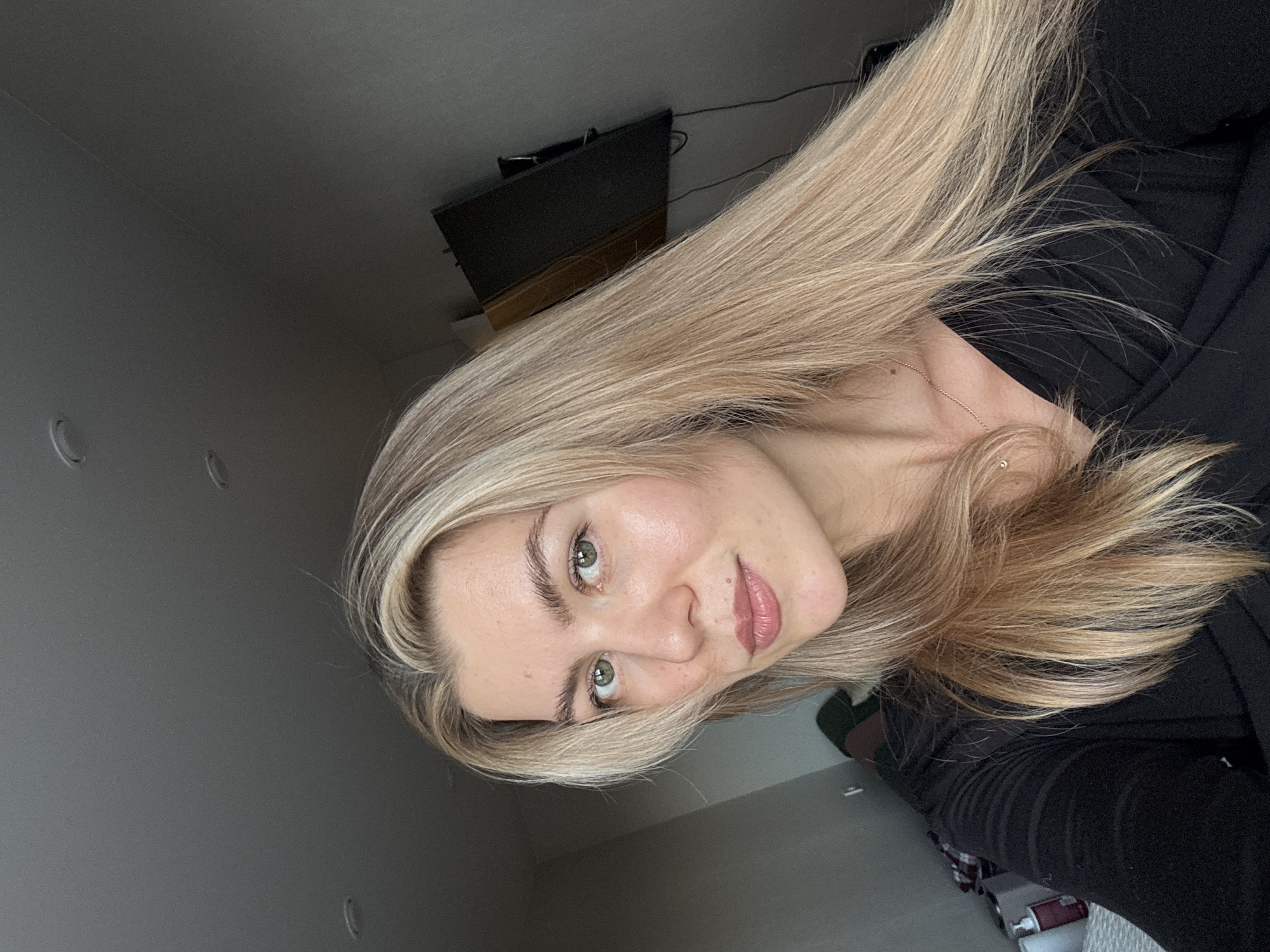 A young woman with blonde hair, fair skin, and light eyes is posing for a selfie in an indoor setting. She is wearing a black top and a delicate gold necklace. Her hair is styled to the side, and she has light makeup on. The background features a wall, a television mounted on the wall, and a glimpse of the floor. The scene is casually lit, suggesting natural lighting from a window. The location is Unknown, Unknown.