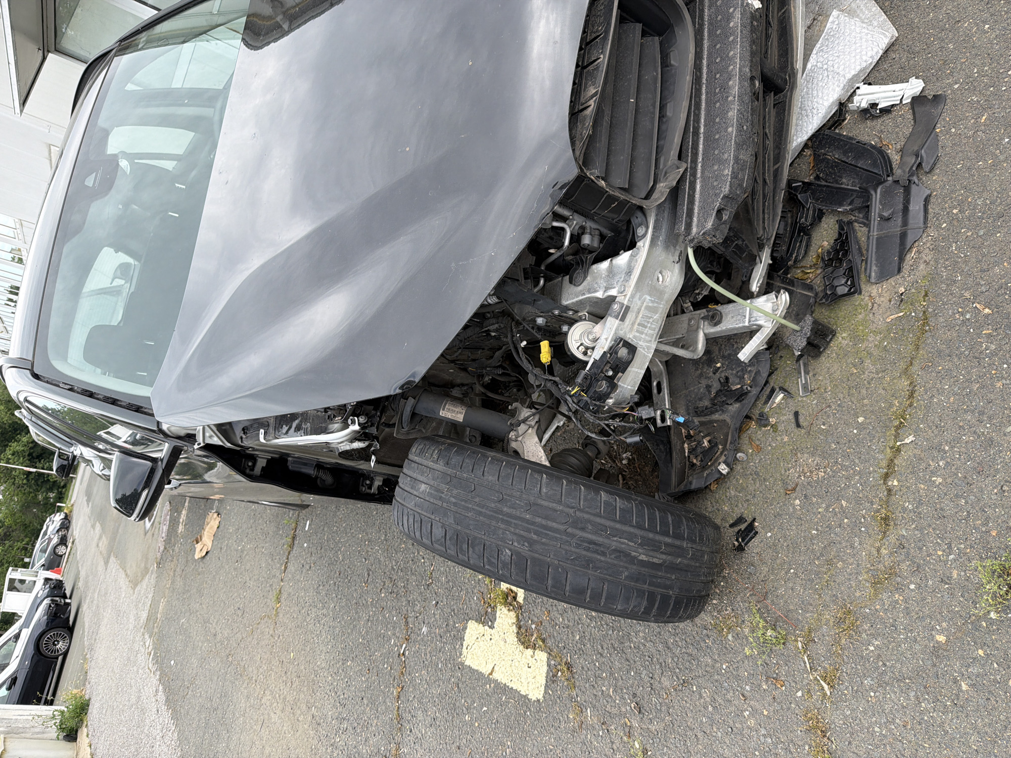 The image shows a severely damaged dark-colored car. The front end of the car is heavily broken and mangled, with the front bumper missing and the internal mechanics exposed. One of the front tires is detached from the car and rests on the asphalt. Debris from the car is scattered on the ground near the front. Other cars are parked in the background, and a building can be seen in the far upper left. The location is a parking area. There is no evidence that the location is in Burgas, Bulgaria. No people are visible.