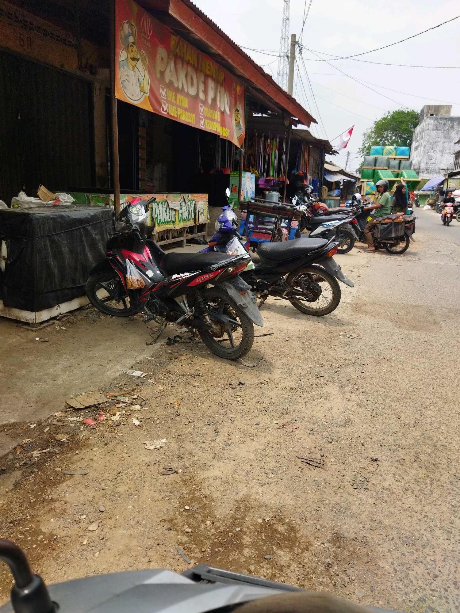 The image depicts a street scene in Banjar Agung, Indonesia, with a focus on a row of small shops or stalls lining the right side of the road. In the foreground, two motorcycles are parked on the dirt and gravel roadside. The motorcycle on the left is red and black, with a plastic bag containing food hanging from its handlebars. The motorcycle on the right is black. Behind the motorcycles, the shops have open fronts with various goods displayed, including cleaning supplies like brooms and mops. A large red and yellow banner hangs above one of the stalls, featuring an illustration of a chef and text in Indonesian, including "PAKBUE PIN" and a list of food items such as "MIE AYAM," "MIE PANGSIT," and "LUCU ONG."
Further down the street, more motorcycles and a few people are visible. One person is riding a motorcycle and appears to be looking towards the viewer. Another individual is seated on a motorcycle with a basket attached to the rear, also facing the street. In the background, there are stacks of goods, possibly building materials or packaged items, covered in green and blue tarps. A tall telecommunications tower is visible in the distance, along with power lines crisscrossing the sky. The overall environment appears to be a busy local market area with a mix of commercial activity and pedestrian traffic. The ground is a mixture of dirt, gravel, and some scattered debris. The sky is overcast.