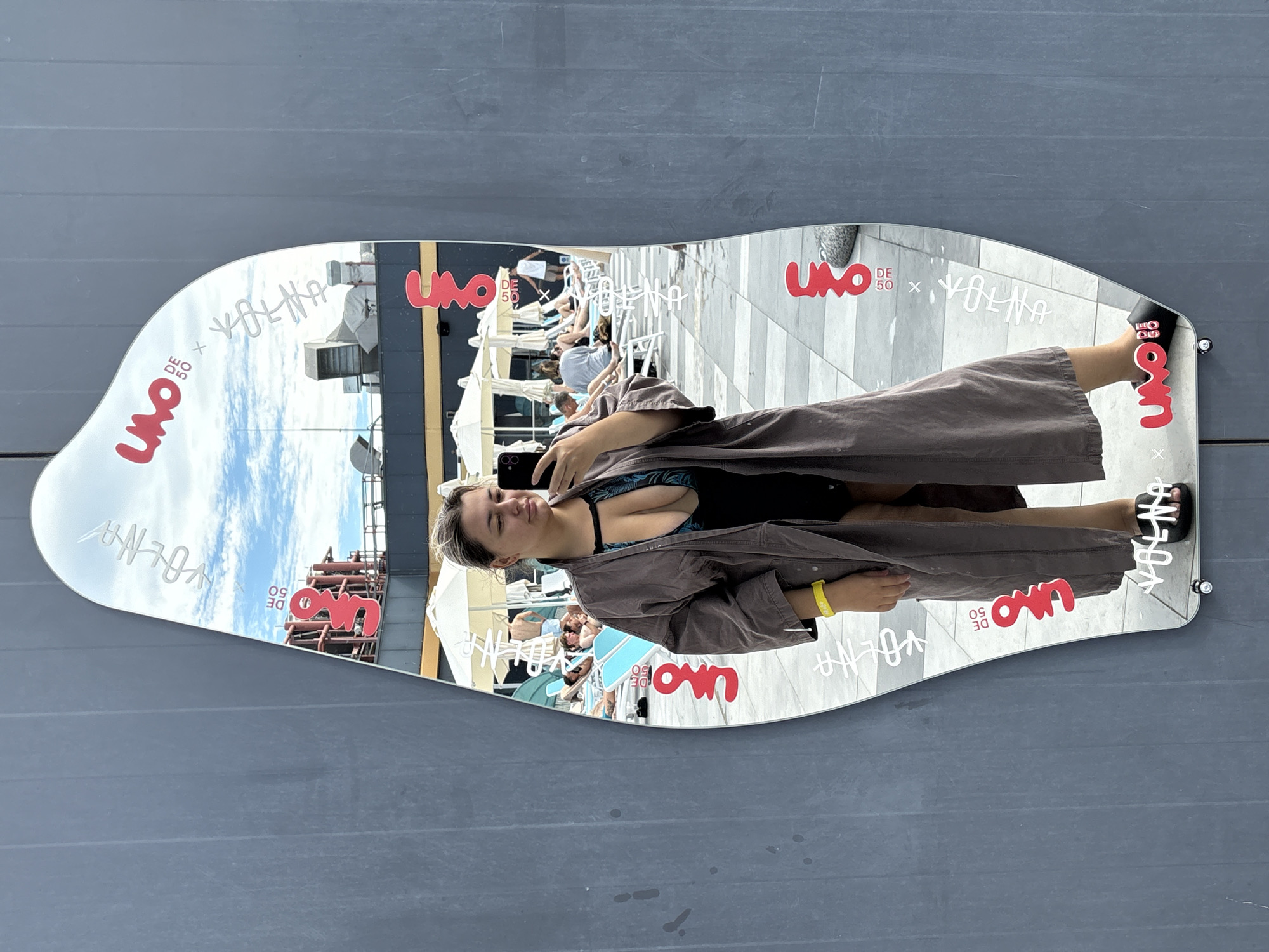A woman is taking a mirror selfie in front of a gray wall. She is wearing a dark brown robe over a black swimsuit with a tropical pattern. She has on black slides. A yellow wristband is visible on her left wrist. The mirror is an irregular shape and has several red and white logos on it. In the reflection of the mirror, a pool area with lounge chairs and umbrellas is visible, along with other people. The scene appears to be outdoors during the daytime with a cloudy sky. The logos in the mirror suggest this location is associated with "UNO DE 50" and "VOLNA". Based on the provided context, the location is the city of Khabarovsk, Russia.