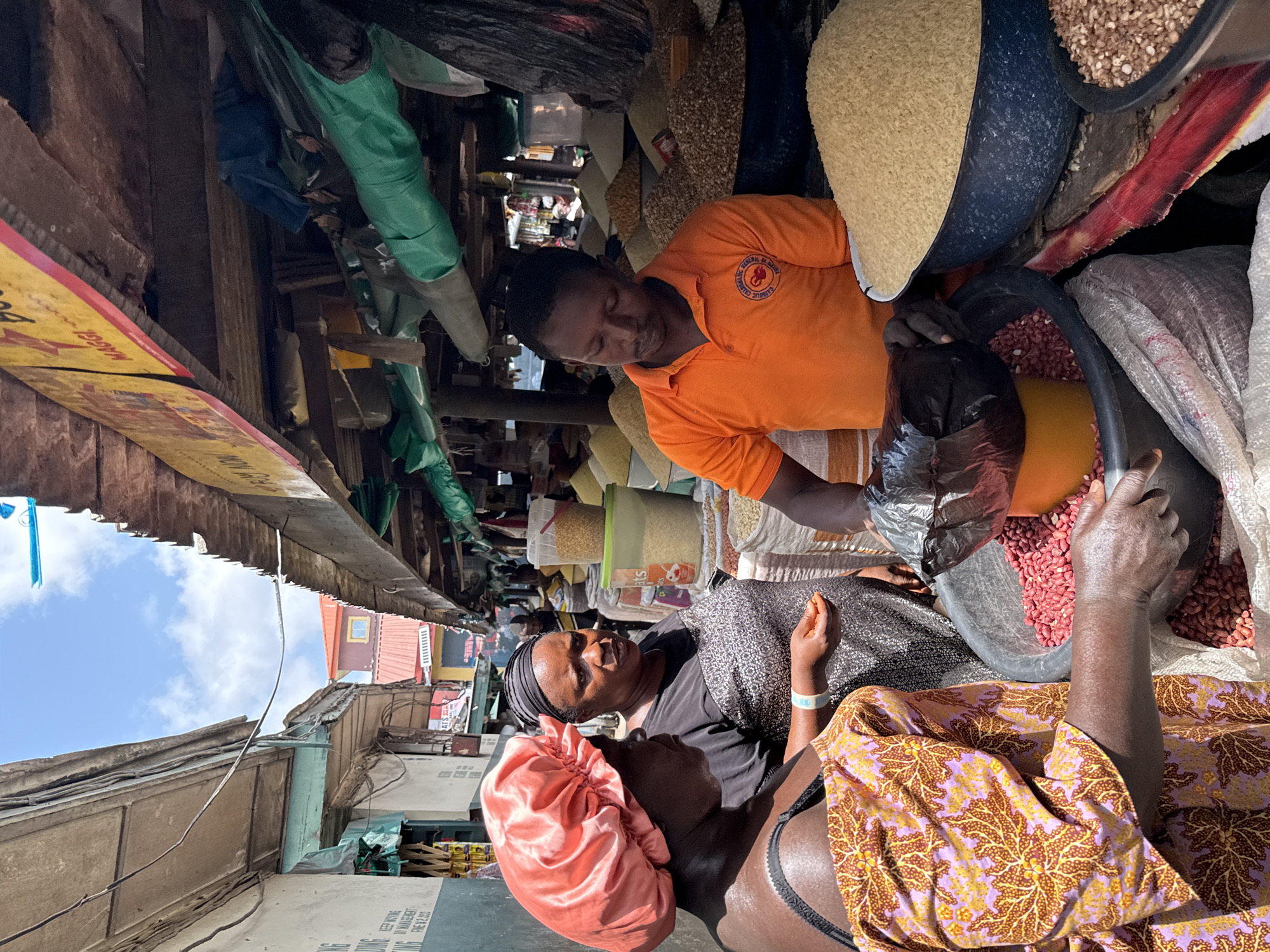 The image captures a vibrant, low-angle view of a bustling market stall in Lekki, Nigeria, under bright daylight with patches of blue sky visible through the makeshift roofing.
Three individuals are prominently featured, engaged in what appears to be a commercial transaction. In the mid-ground, a person wearing a bright orange short-sleeved polo shirt with a circular logo on the left chest (partially visible, reading "CAMPUS CRUSADERS OF CHRIST") is leaning over, handling goods – specifically, scooping red beans from a large basin into a black plastic bag. This person is surrounded by other large basins overflowing with various dry food items, including light-colored grains (possibly rice or maize) and more red beans.
To the left and foreground, two women observe or participate. One woman, wearing a black headscarf and a dark patterned top, looks towards the person in orange. She has a blue and white wristband on her right wrist. Partially visible in the immediate foreground is another woman, adorned in a vibrant purple, yellow, and brown patterned garment and a pink satin head covering.
The market setting is characterized by overhead wooden structures, beams, and draped green fabrics, creating a shaded but open environment. A prominent yellow and red sign, partially visible on the left, displays the text "MAGGI." The overall scene depicts a lively, crowded market focused on the sale of staple food items.