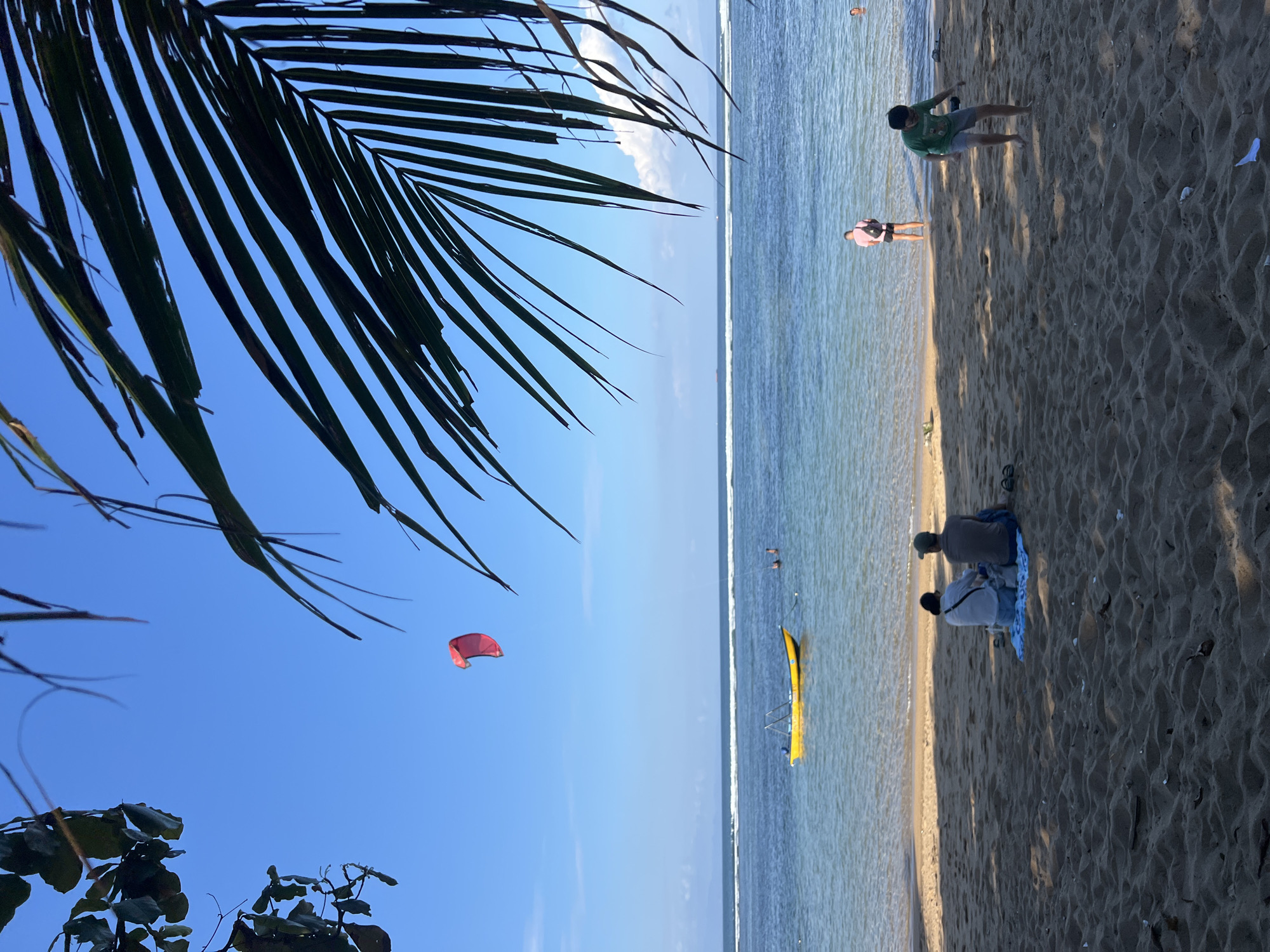 In Denpasar, Indonesia, on a sandy beach, a bright yellow boat is anchored in the shallow ocean. A person is visible in the water behind the boat. In the distance, a red kite flies in the bright blue sky above the ocean. On the beach, two individuals sit facing away from the camera, while a young person in a green t-shirt walks towards the water. The scene is framed by the fronds of a palm tree in the foreground.