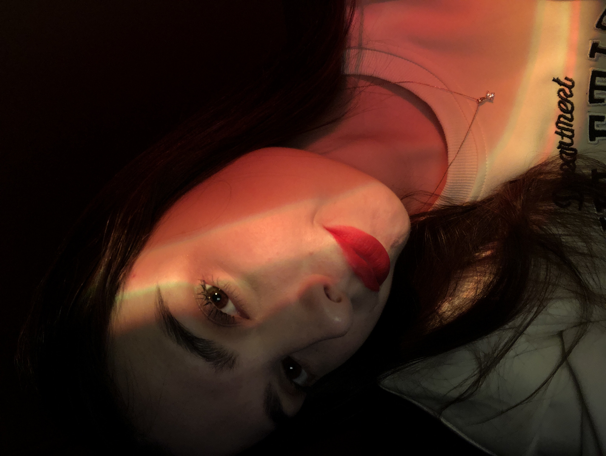 A portrait photograph captures a young woman in a dimly lit indoor setting. She is positioned as if reclining, with her head tilted, and her dark hair cascades around her. A prominent diagonal beam of red and subtle rainbow-colored light cuts across her face, illuminating her left eye, nose, and vibrant red lips. Her gaze is directed towards the viewer, conveying a neutral or slightly contemplative expression.
She wears a light-colored, possibly white or very light grey, crew-neck sweatshirt. Around her neck is a delicate necklace featuring a small, clear stone pendant. Part of an embroidered word is visible on her sweatshirt, reading "Department". The background is dark and indistinct, creating a sense of intimacy and dramatic contrast with the illuminated features. The lighting suggests the image was taken in the evening or at night. There are no specific visual cues within the image itself to indicate the Noviy, Russia, location context.
