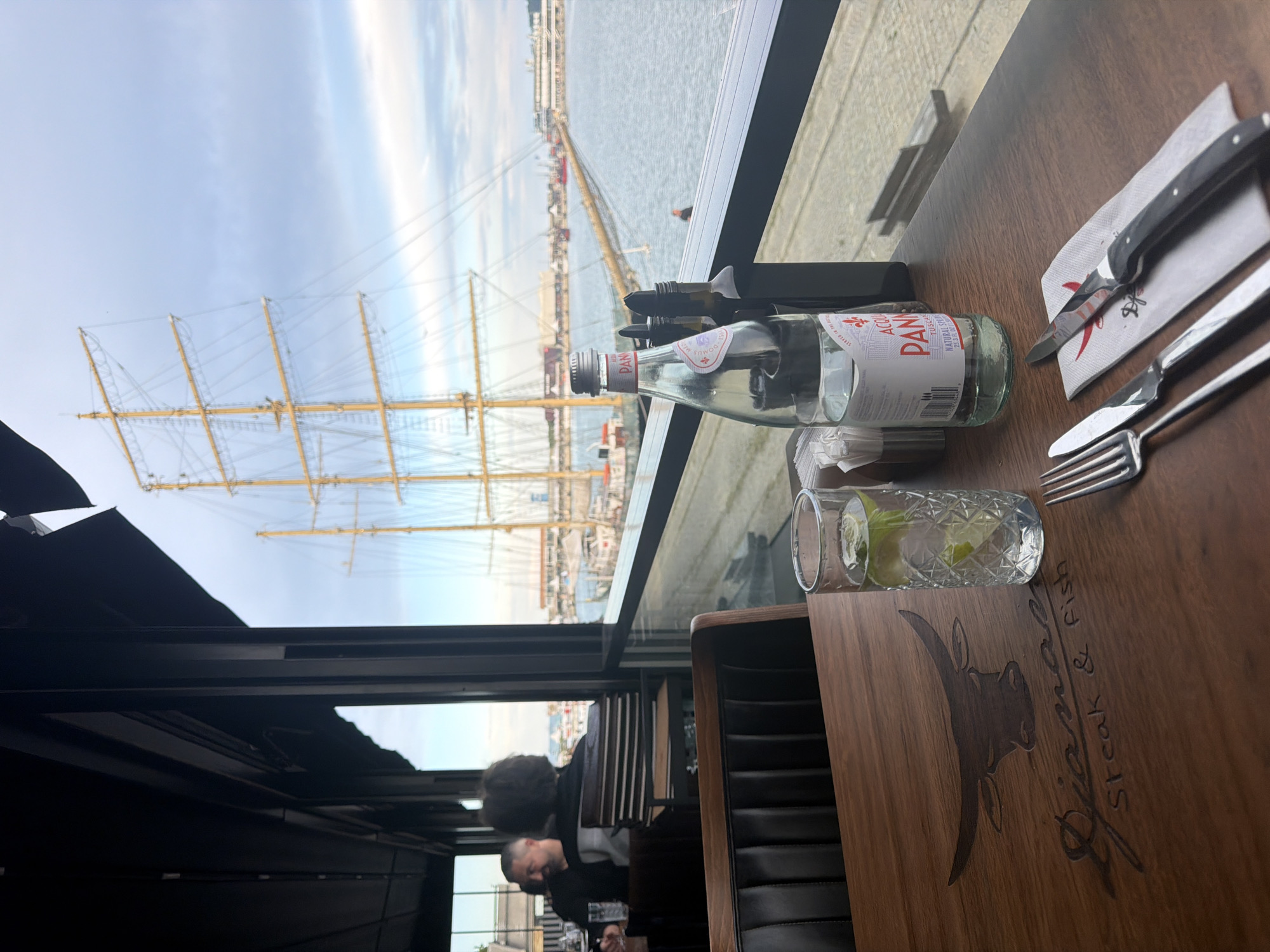 This media file shows a table setting in a restaurant with a view of a docked sailing ship. The table is set with a bottle of San Pellegrino water, a glass with ice and lime, a knife, and forks. The restaurant appears to have a dark wood interior. In the background, through a large window, a tall sailing ship with multiple masts and sails is visible, docked in a harbor. The sky is cloudy with patches of blue. The scene suggests a daytime meal. The visible text on the bottle reads "San Pellegrino Acqua Panna." On the wooden table, a logo is visible which appears to read "Gjiana Steak & Fish." The setting is likely a waterfront restaurant in Varna, Bulgaria, given the context. There are no explicit emotions conveyed, but the scene is peaceful and observational.