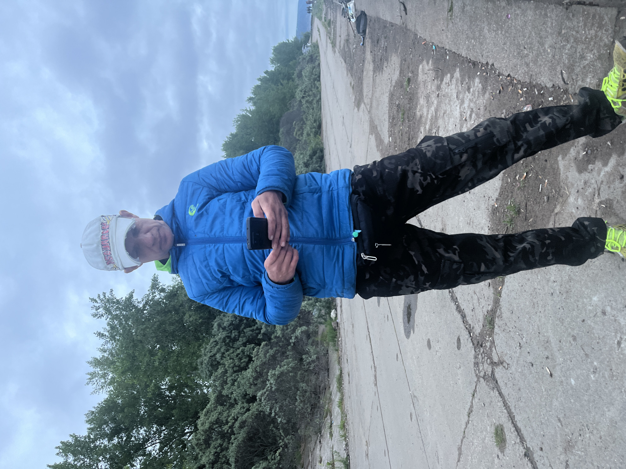 An adult male stands centrally on a cracked concrete path in Chaikovsky, Russia, observed from a low angle. He wears a white baseball cap, a blue puffer jacket with a visible green lining at the collar, dark camouflage cargo pants, and bright lime green and yellow athletic shoes. He holds a black smartphone horizontally with both hands, directing his gaze downward at the screen. The path, marked by cracks, dirt, and small puddles, extends into the background, curving slightly to the right. Dense green foliage, comprising trees and bushes, borders the path on both sides. In the mid-ground, a vehicle is partially visible behind the trees on the right. Further down the path on the right, several fishing rods and a black bag are discarded on the ground. A group of indistinct figures and a faint horizon line are visible in the far distance. The sky is overcast with uniform gray clouds.