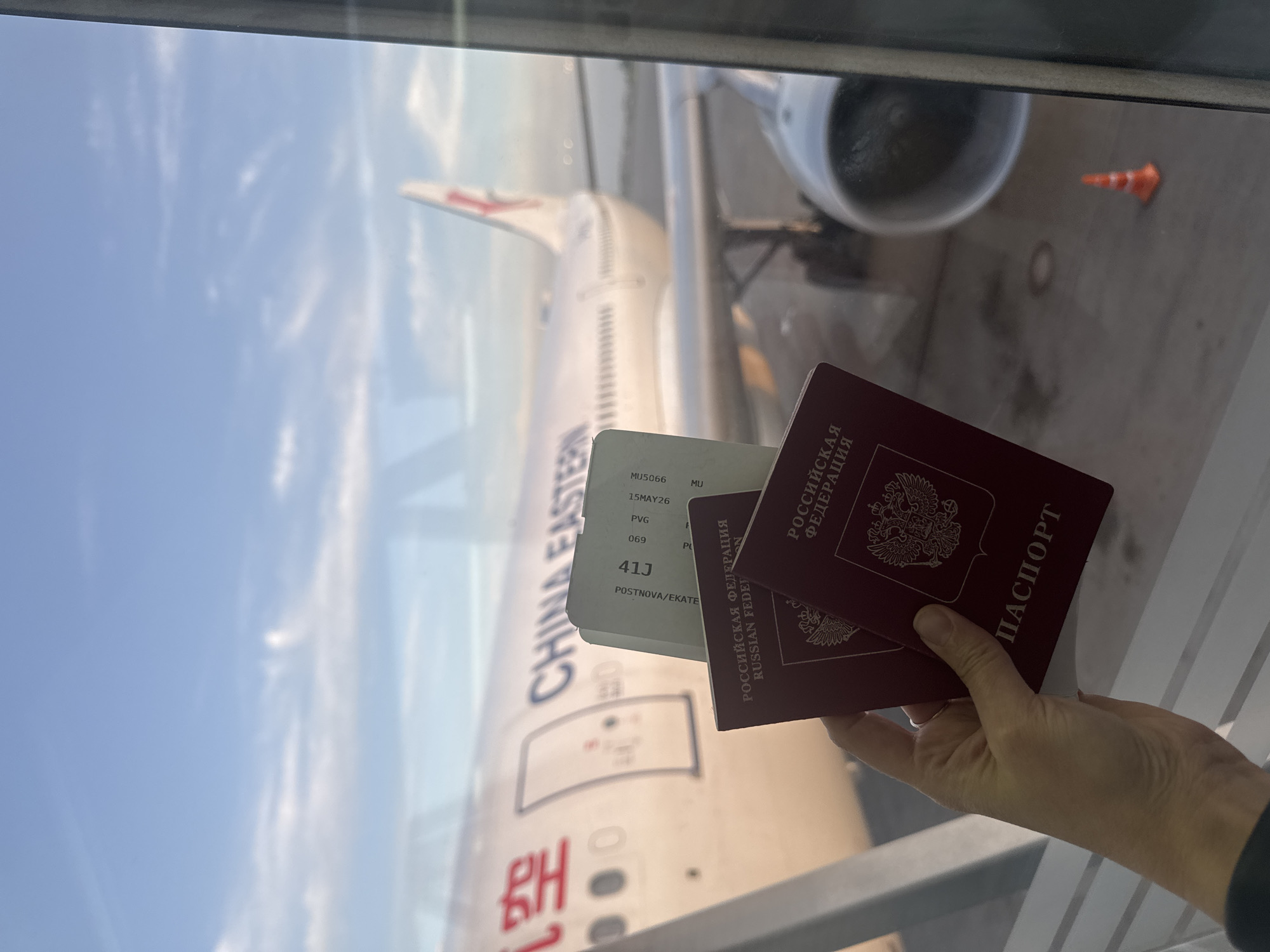 Here's a detailed description of the image:
**General Description:** The image shows a person holding two Russian passports and a boarding pass in front of an airplane window. The aircraft visible outside is a China Eastern Airlines plane.
**Specific Details:**
* **Passports:** Two maroon-colored Russian passports are visible. The inscription "РОССИЙСКАЯ ФЕДЕРАЦИЯ" (RUSSIAN FEDERATION) is clearly visible on the cover of one, along with the word "ПАСПОРТ" (PASSPORT).
* **Boarding Pass:** A boarding pass is partially visible, showing details such as the flight number "MU5066", the date "15MAY26", departure airport "PVG" (Shanghai Pudong), the destination airport, a seat number "41J", and the name "POSTNOVA/EKATE".
* **Aircraft:** The exterior of a China Eastern Airlines aircraft is visible through the window. The airline's name and logo are clearly displayed on the fuselage.
* **Environment:** The scene appears to be inside an airport terminal, near a gate. The window suggests the person is inside the terminal, looking out at the plane. Part of the tarmac and possibly some airport equipment are visible outside.
* **Person:** The hand holding the passport and boarding pass belongs to a person whose features are otherwise obscured.
* **Location:** Given the text "POSTNOVA/EKATE" on the boarding pass, it's possible that the destination is in the Ekaterinburg area. However, the origin point is clearly Shanghai Pudong airport. It's not possible to state that the location is in Bol'shie Kabany, Russia, as that does not appear to be a location related to the details in the photo.