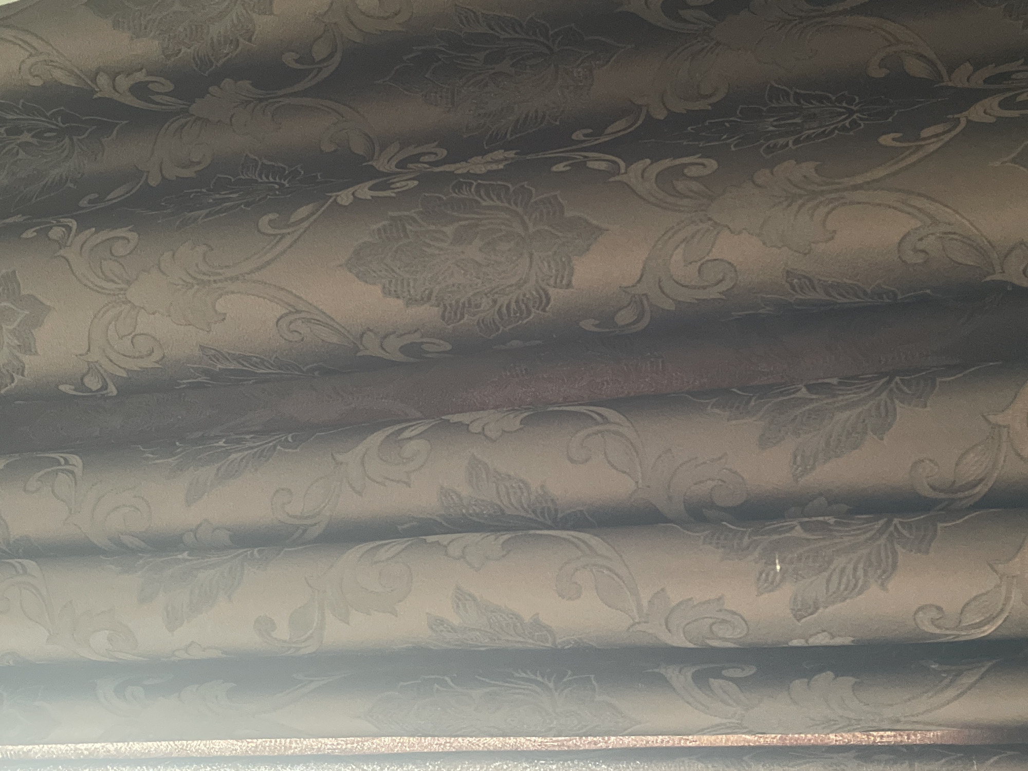 A close-up shot shows a brown damask curtain with a floral pattern. The fabric has a subtle sheen, and the folds of the curtain create a play of light and shadow, highlighting the raised patterns. The curtain appears to be hanging in a room, possibly in a residential or hotel setting. The image does not provide any specific information to determine its location in Tallinn, Estonia.