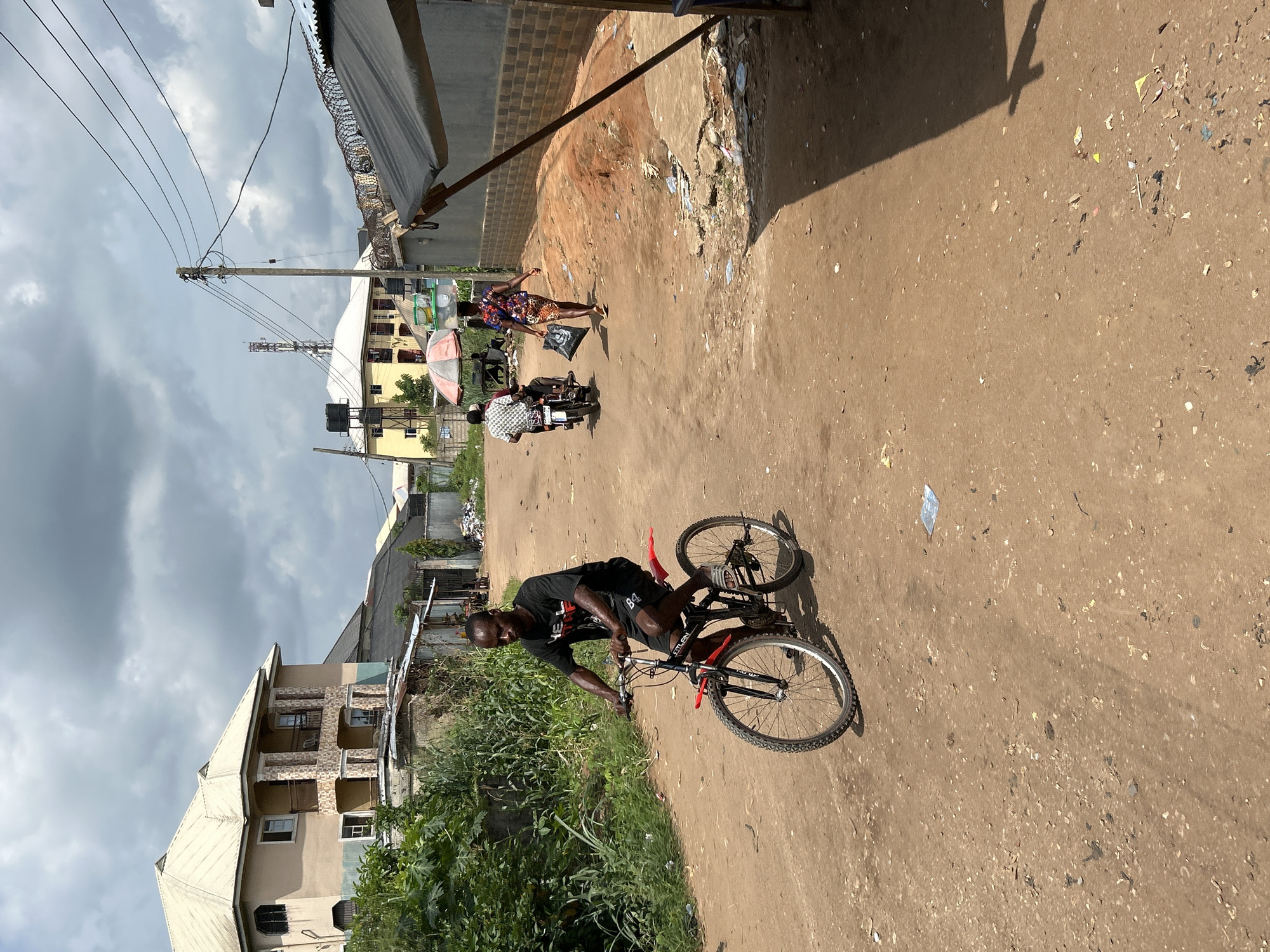 The image shows a man riding a bicycle on a dirt road in Ihiagwa, Nigeria. The road appears to be unpaved, with scattered debris and garbage. Buildings are visible in the background, including residential structures and shops. Other people are present on the street, including a woman carrying a bag and two individuals on a motorcycle. An overhead power line stretches across the scene, and the sky is overcast.