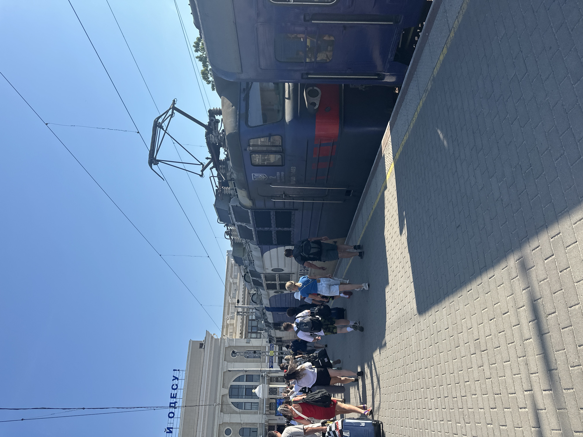 The image depicts a sunny day at the Odesa railway station in Odesa, Ukraine. The station building, a grand structure with classical architectural elements, is visible in the background. A train, painted in blue and white, is parked at the platform. Several people, appearing to be passengers, are walking along the platform, some with luggage. The platform itself is paved with brick pavers, and a yellow line demarcates the edge. Overhead, the blue sky is crisscrossed with power lines for the electric train. The overall scene conveys a sense of travel and activity at a bustling transportation hub.