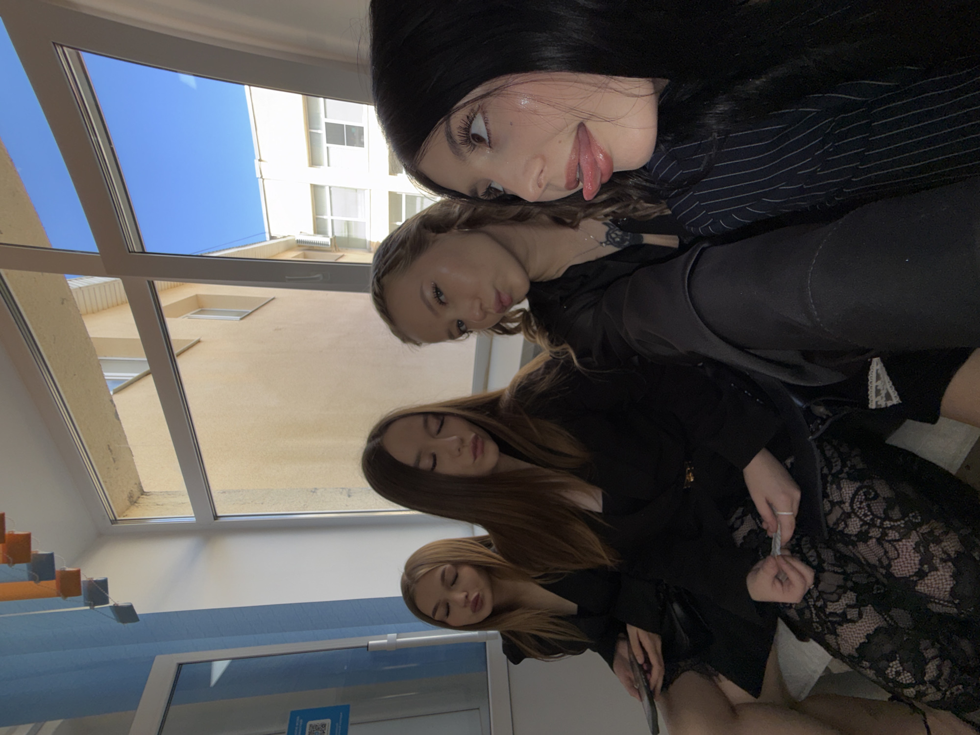 Four young women are seated in front of a window with a view of buildings and a blue sky. The woman in the foreground, on the right, is looking to the right and has her tongue sticking out. She is wearing a black pinstripe jacket. To her left, a woman with blonde hair is looking towards the camera with her lips pursed. Next to her, a woman with long brown hair is also looking down with her lips pursed. The woman on the far left, also with long brown hair, is looking down with her lips pursed and holding a phone in her left hand. The women are dressed in dark clothing, with two of them wearing black blazers and lace skirts. The environment appears to be indoors, possibly a classroom or common area, with a window and part of a blue wall visible on the left.