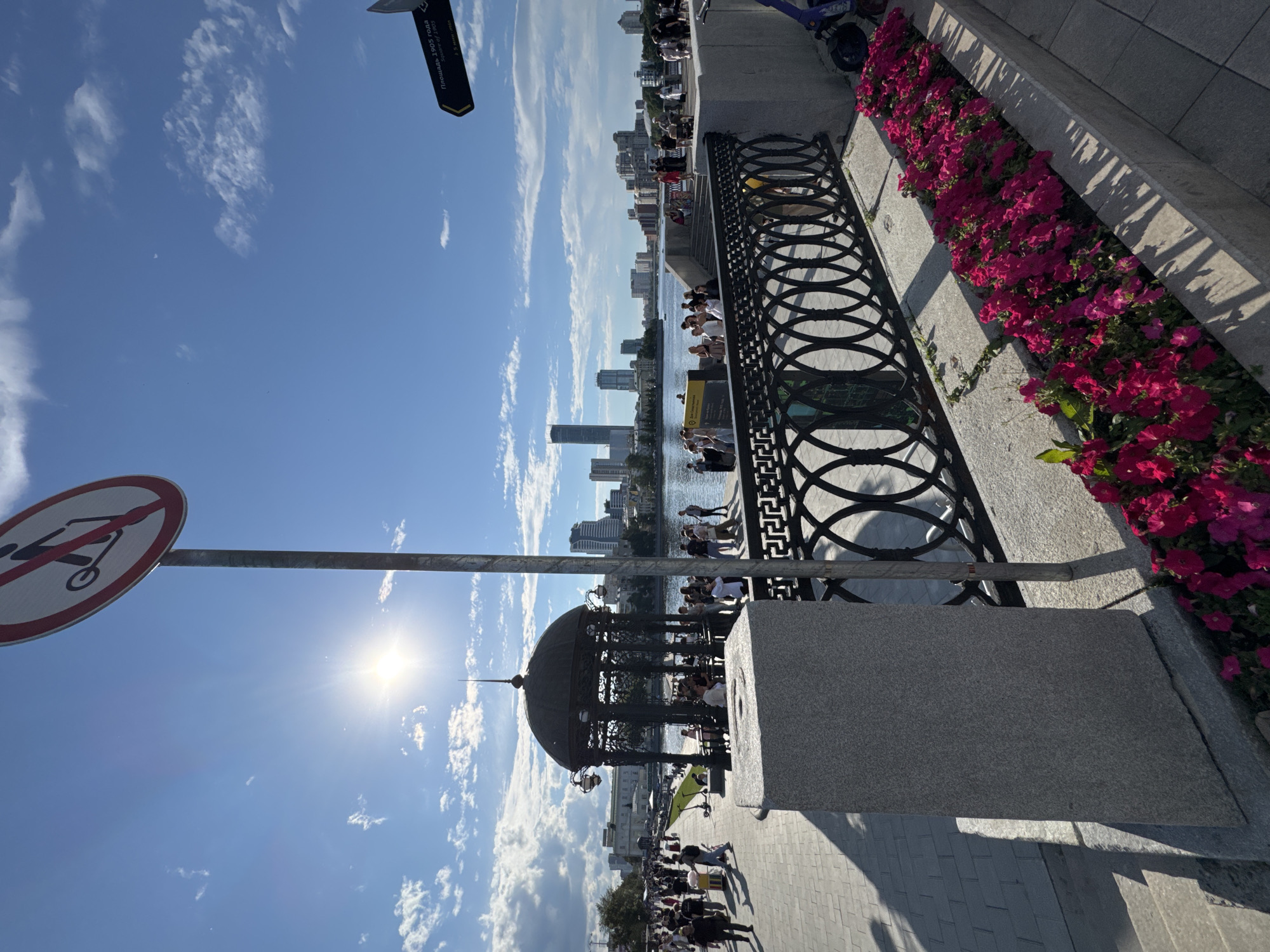 The image depicts a sunny day in Yekaterinburg, Russia, likely in the late morning or early afternoon due to the bright sunlight and clear blue sky with scattered clouds. The scene is a waterfront promenade, bustling with people enjoying the outdoors.
In the foreground, a circular sign with a red border prohibits the use of scooters. A metal railing with an intricate circular pattern runs along the edge of the promenade, separating it from the water. To the right, a planter overflows with vibrant pink flowers, casting long shadows onto the paved walkway. Further along the promenade, a distinctive, ornate dark-colored gazebo stands, providing a shaded gathering spot.
The background reveals the Yekaterinburg cityscape with modern high-rise buildings lining the opposite bank of the river or canal. People are scattered throughout the scene, some walking, others sitting or standing near the water's edge. The overall atmosphere appears relaxed and lively, suggesting a public space actively being utilized by residents and visitors on a pleasant day. A blue electric scooter is visible near the top of the frame. A black sign with Cyrillic text is attached to a pole, partially obscured by the sky.