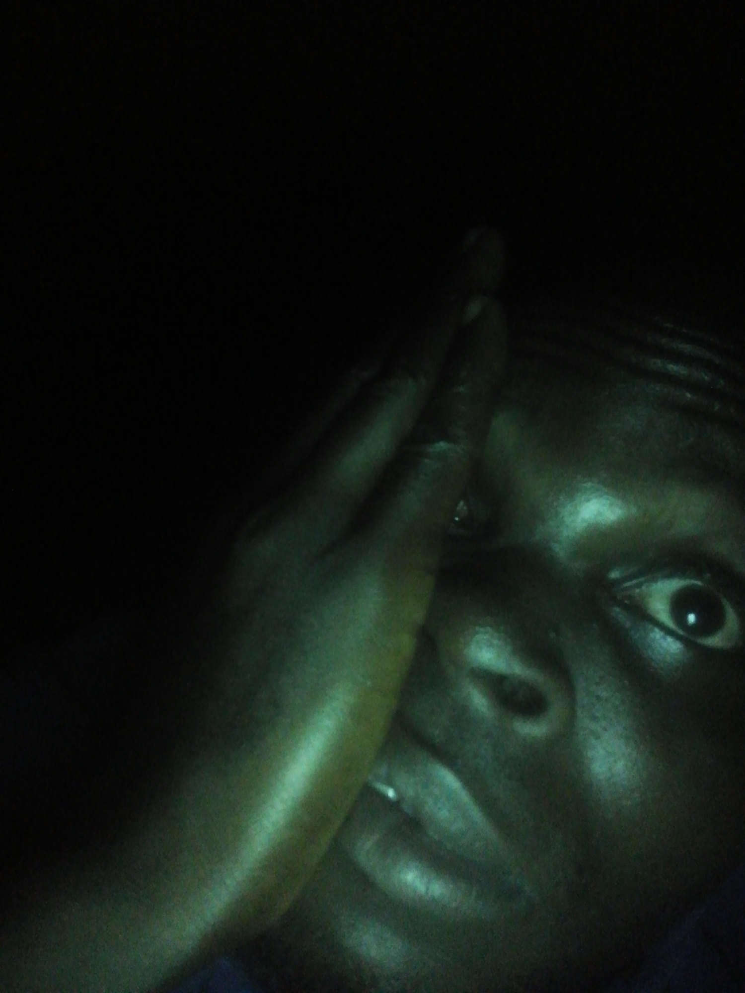 A close-up, low-angle shot features a dark-skinned individual obscuring part of their face with a hand. The lighting is poor, creating deep shadows and emphasizing the texture of the skin. The person's eye is visible, looking directly at the viewer. The background is black.