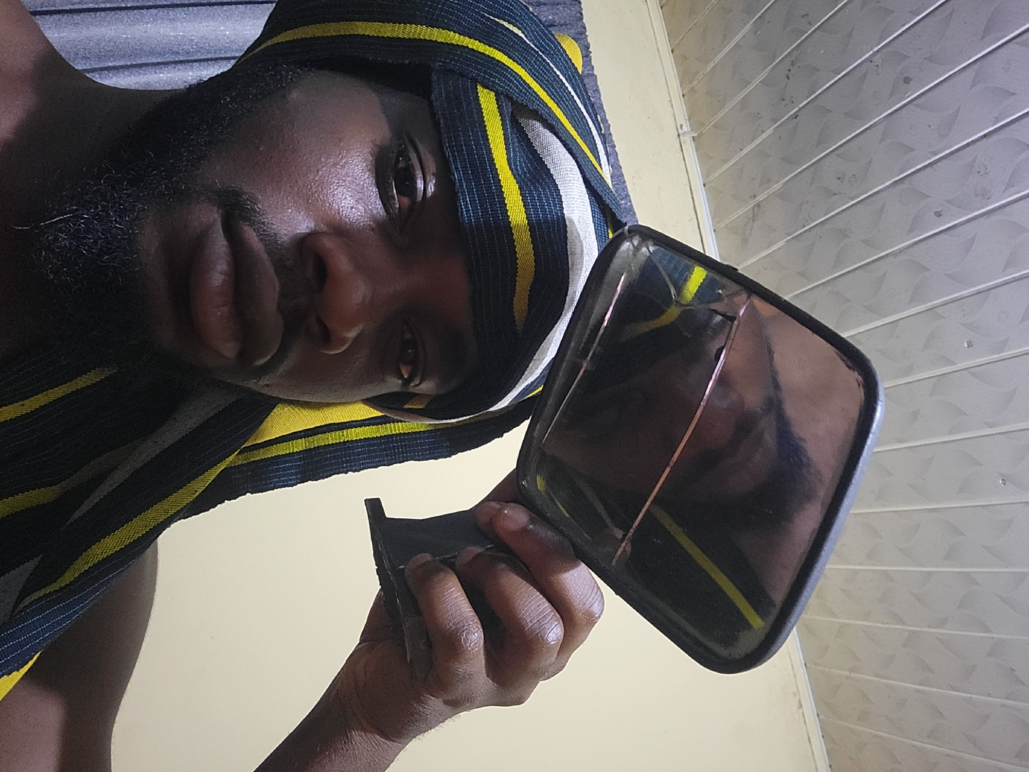 A dark-skinned male subject, estimated to be between 20 and 35 years old, is depicted indoors in Jos, Nigeria. He has a short, dark beard and mustache, and his skin appears to have a slight sheen. He is wearing a headwrap made of a striped fabric featuring dark blue/black, yellow, and white/light grey patterns. The subject holds a small, rectangular handheld mirror with a dark frame and handle in his right hand, angled upwards. The mirror's reflective surface shows a distorted view of a light-colored ceiling. The background features a ceiling constructed from multiple light grey or white PVC panels arranged in strips, exhibiting a subtle repeating texture. A plain, light-colored wall is visible behind the subject, with a partial view of a darker, possibly curtained, surface on the left.