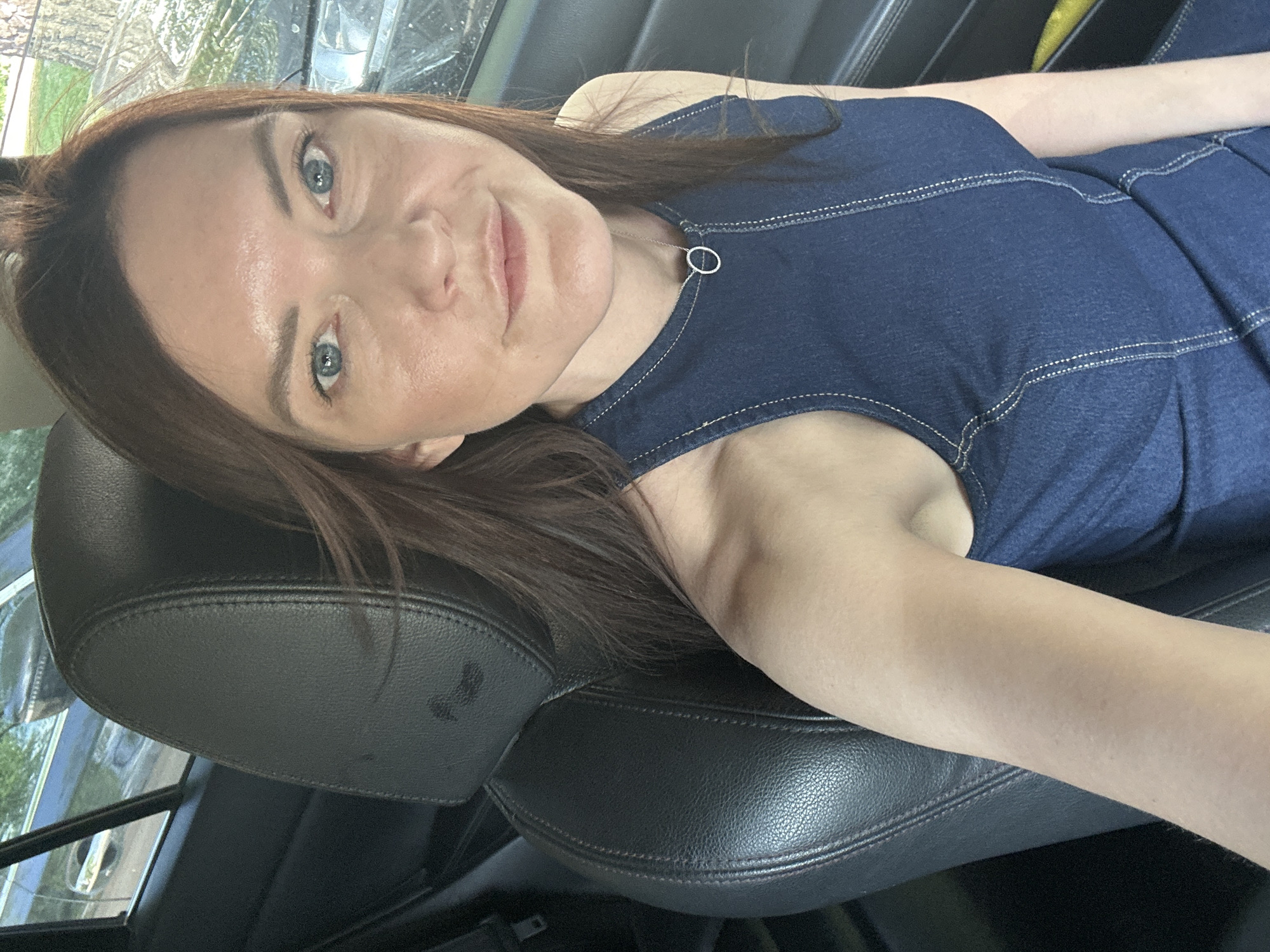 A woman with light skin and auburn hair is reclining in a black leather car seat. She is wearing a blue denim-like dress with white stitching around the neckline and armholes, and a necklace with a circular pendant. The setting appears to be inside a car, with a view of a residential area visible through the window. The image provides no indication that the location is Chaikovsky, Russia.