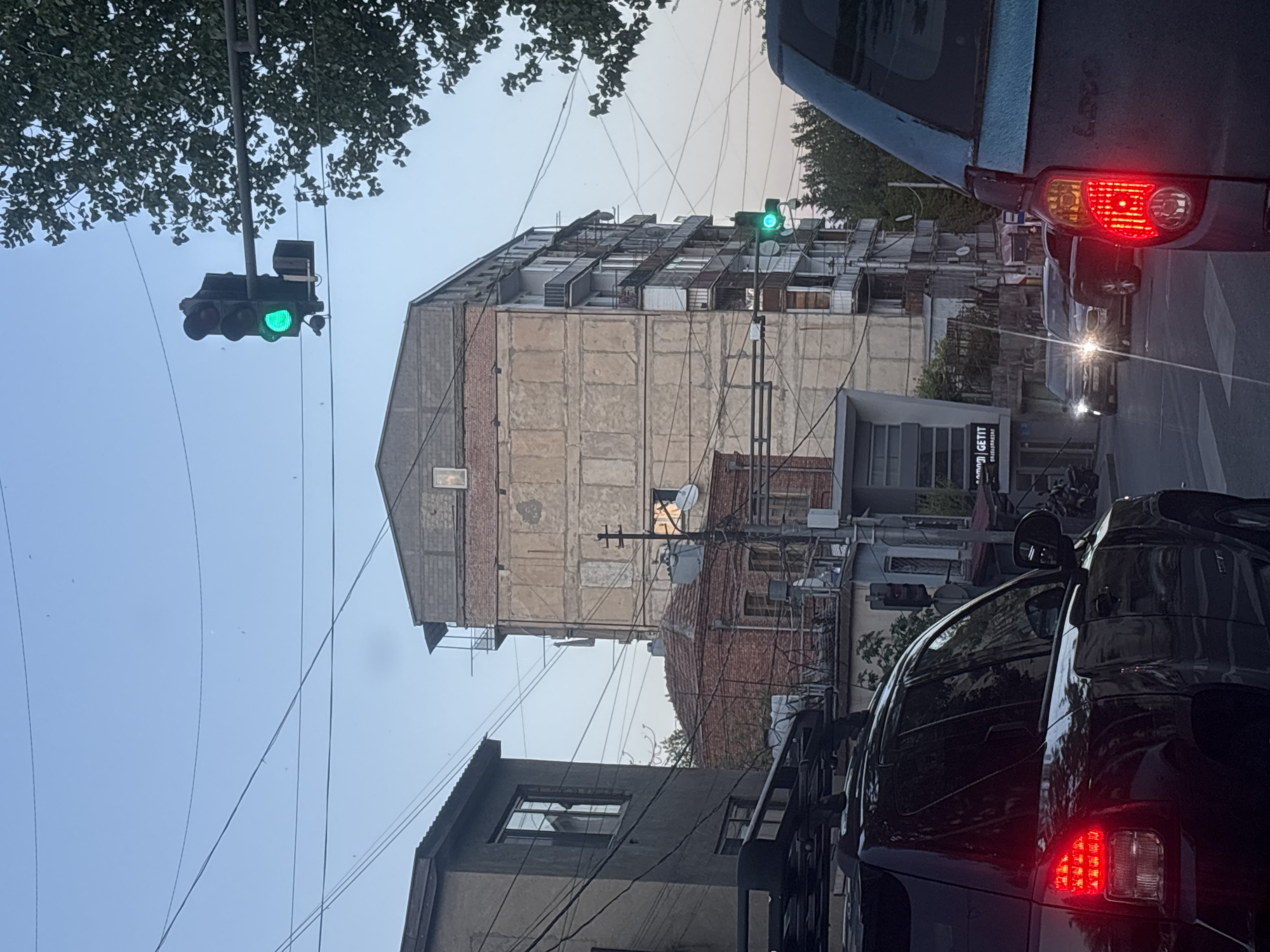 The image captures an urban street scene from a vehicle's perspective, looking forward while stopped in traffic. The original image is rotated 90 degrees clockwise, showing a vertical horizon.
In the foreground, the rear of a blue car with illuminated red taillights is prominent on the right, indicating it is stopped. The vehicle from which the photo is taken is also visible on the right, with its side mirror and parts of its body. A traffic light, suspended by wires, hangs above the street on the left, displaying a green signal. Another green traffic light is visible further down the road, to the right of the central building.
The midground is dominated by a tall, multi-story building with a somewhat weathered appearance. Its facade features large, light-colored rectangular concrete panels and sections of exposed brickwork. The building's rooftop is distinct, covered with numerous interconnected structures that resemble vents or small rooftop units. To the right of this building, another structure with a brick facade and various cables and wires attached is visible. Below it, on a darker building, a commercial sign with the partial text "GET FIT" and "GETTI" is discernible, along with a partial phone number.
The sky is clear and bright, suggesting either early morning or late afternoon light, with soft shadows. Numerous power lines and utility cables crisscross the frame against the light sky. Further down the street, other vehicles are visible, some with illuminated headlights. No people are clearly visible. The text "LOGOS" is faintly visible on the rear of the blue car in front. The architectural style and dense overhead wiring are consistent with an urban setting like Tbilisi, Georgia.