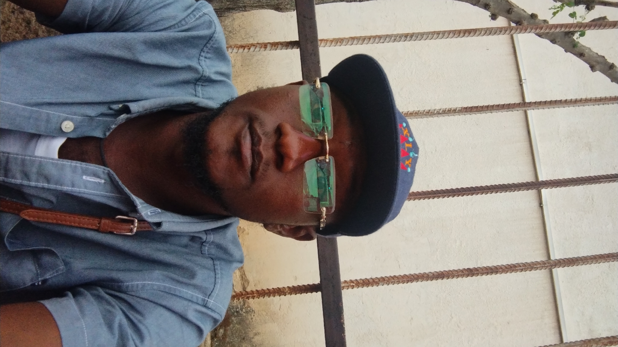 The image is a portrait of a dark-skinned man in a close-up shot, looking directly at the camera. He is wearing a dark blue baseball cap with a colorful embroidered logo that appears to be "X-RAY." He has on rimless glasses with light green tinted lenses. His clothing consists of a light blue, short-sleeved button-down shirt, worn over a white undershirt. A thin brown strap, possibly from a bag, is visible crossing his chest. He has a goatee and a short beard. The background features a textured, light-colored wall and vertical rusty metal bars. The lighting suggests it is daytime, with some shadows indicating an outdoor setting. The man appears to be outdoors, possibly in a courtyard or alleyway, given the wall and metal bars. The style of the building materials and the presence of the tree on the left suggest a location within Nigeria. Specifically, the context provided by the user points to Jos, Nigeria, which is known for its diverse architecture and urban environments.