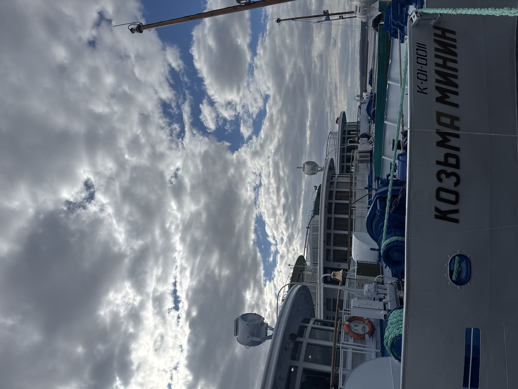 The image shows a section of the river passenger ship "Kozma Minin," as it is painted on its side in Cyrillic, along the Kama River. The ship is a light gray color, and some blue mooring lines are visible. The sky above is filled with fluffy clouds. The photo appears to be taken in Chaikovsky, Russia, a city located along the Kama River.