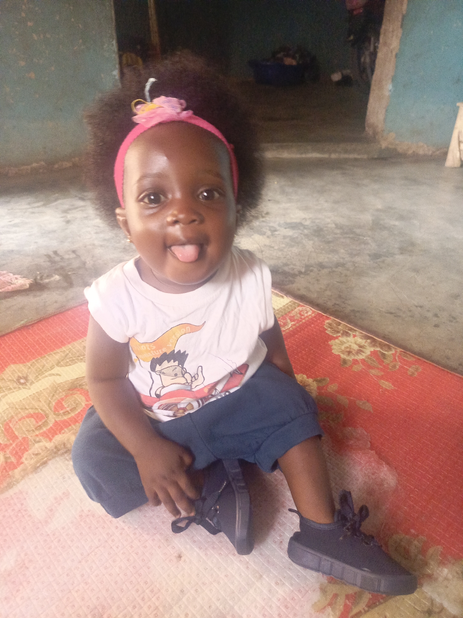 A young child is sitting on a patterned rug indoors. The child is wearing a pink headband with a flower, a white t-shirt with a cartoon graphic, dark blue shorts, and black shoes. The child is sticking their tongue out and looking at the camera. The room has a concrete floor, and there are items in the background, suggesting a domestic setting. The location is Unknown, Unknown.