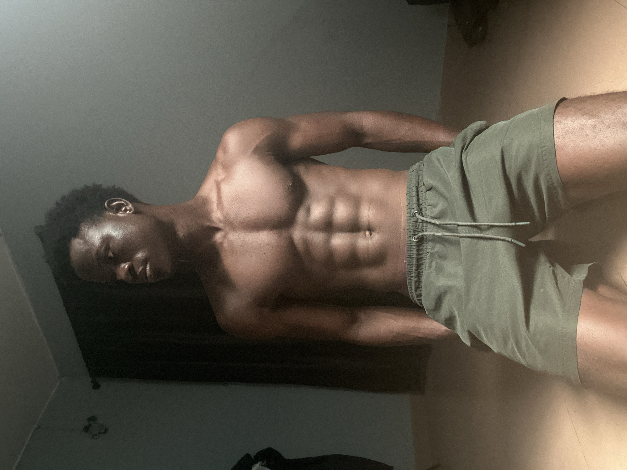The image shows a young Black male with a muscular physique kneeling indoors. He is shirtless, wearing green shorts. He is looking to his left, with his right hand in his pocket and his left arm resting on his left leg. The background consists of a dark curtain and a light-colored wall. The floor appears to be tiled. The lighting is dim, with a strong light source coming from the right, casting shadows. This scene is likely located in Badagry, Nigeria, based on the provided information, though there are no specific visual cues in the image to confirm this location.