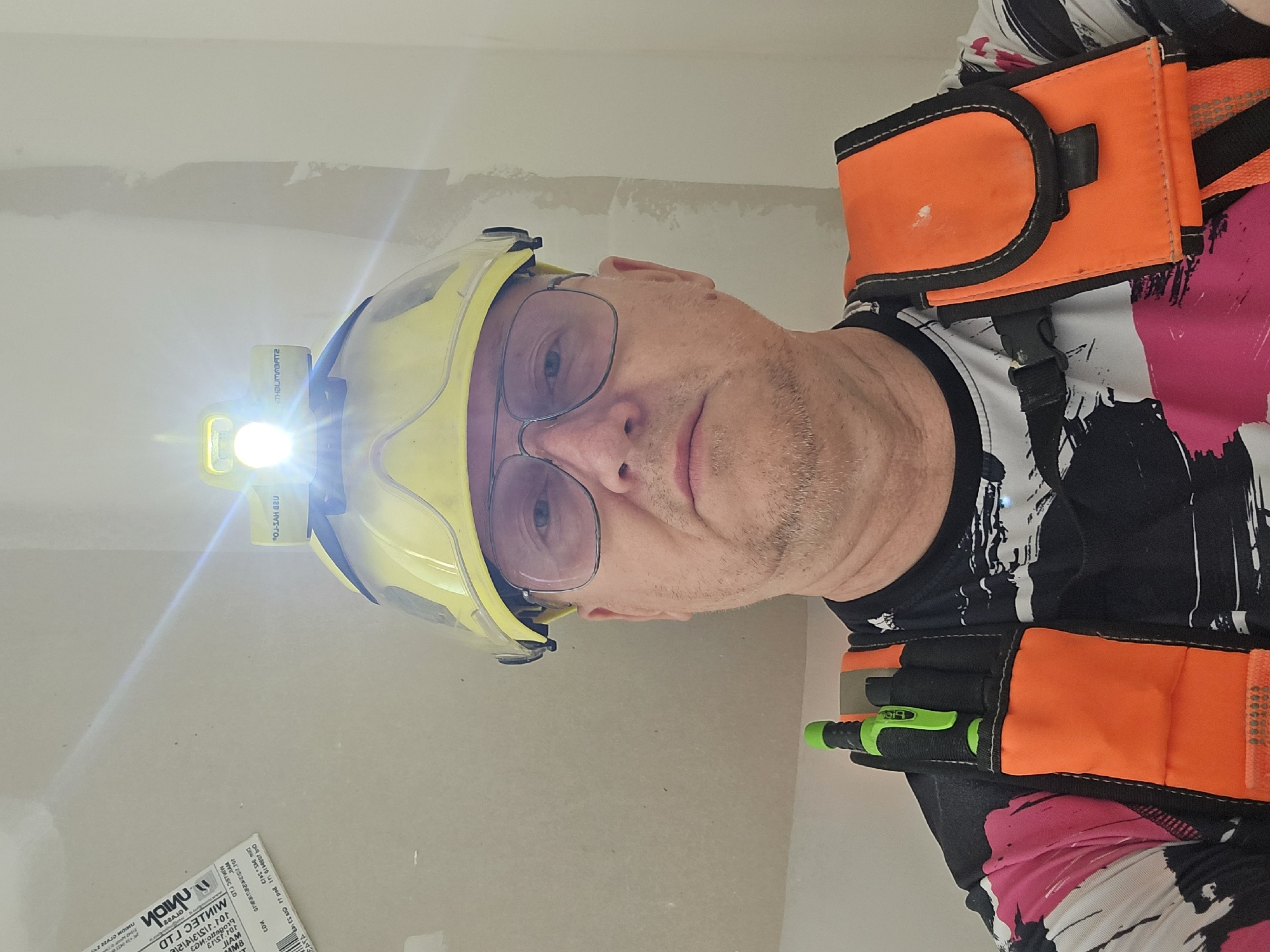 A man wearing a yellow hard hat with a headlamp, glasses, and an orange and black vest is indoors with drywall visible in the background. The headlamp is illuminated. The man is wearing a black and white patterned shirt with pink accents. There are tools in the vest. Text related to MINTEC LTD and THELUMABATZ is visible. No indication of Tel-Aviv, Israel is visible.