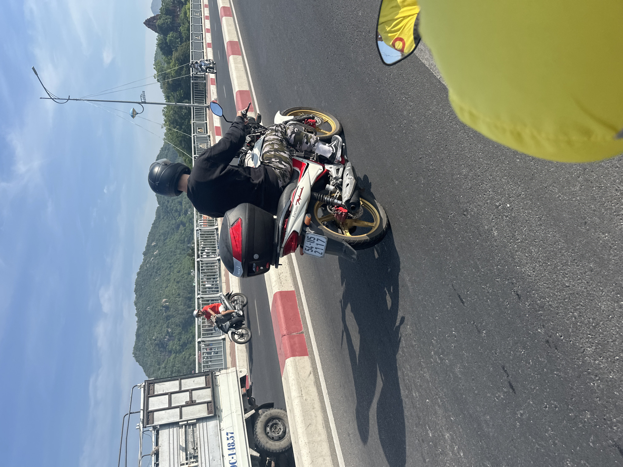 The image depicts a daytime scene on a bridge in Bac Nha Trang Ward, Vietnam, likely during daylight hours given the clear sky and bright lighting. The foreground is dominated by the perspective from a moving vehicle, with a portion of a bright yellow garment and a side mirror with a yellow reflection visible. A motorcycle rider in a black helmet, black long-sleeved shirt, and camouflage pants is the primary subject, riding a white and red motorcycle equipped with a black storage box on the back. The motorcycle has a license plate reading "54-U5 2117". In the middle ground, another motorcycle with a rider in a red shirt and white helmet is visible, along with a white truck to the left. The bridge is lined with a white and red curb, and beyond that, a metal railing. The background features lush green mountains and trees under a blue sky with scattered clouds. The road appears to be asphalt, and the overall impression is of a typical transportation scene on a bridge in Vietnam.