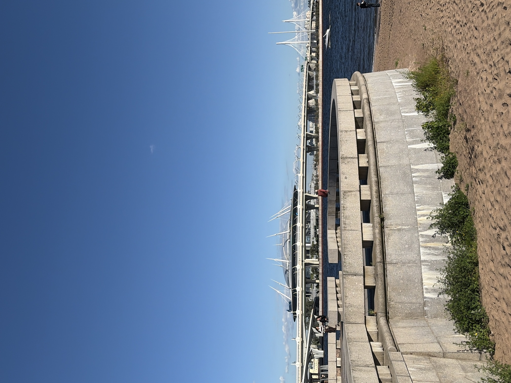 The image shows a scenic view of Saint Petersburg, Russia, on a clear, sunny day. The foreground features a sandy beach and a curved stone embankment with steps, leading down to the water. Lush green bushes and some wildflowers grow along the embankment.
In the middle ground, a long, elevated walkway with a railing runs parallel to the water. Several people are visible walking on this walkway, and one person is sitting on the edge. Beyond the walkway, the blue water of a bay or canal stretches out, reflecting the bright sky. A white boat is moored in the water.
In the background, a distinctive modern bridge with a unique cable-stayed design spans the water. The bridge is a prominent landmark, with its white, angular towers and supporting cables clearly visible against the blue sky. Buildings and other structures can be seen on the far side of the water, hinting at the city's urban landscape.
The overall atmosphere is peaceful and pleasant, suggesting a leisure activity like a stroll along the waterfront. The clear blue sky indicates fair weather, likely midday or early afternoon given the bright lighting. The scene captures a blend of natural elements, urban architecture, and human activity. No visible text is present in the image.