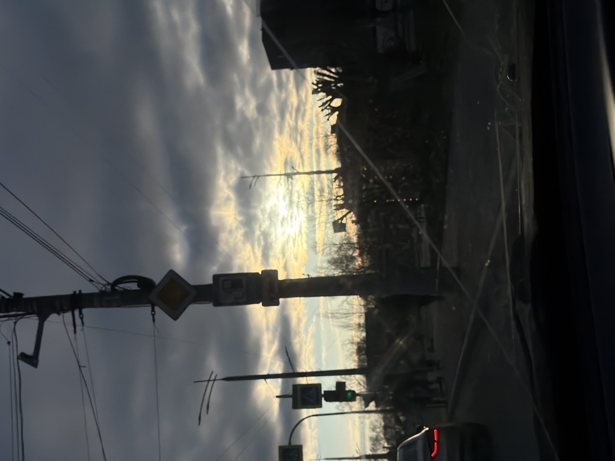 The image, taken from inside a vehicle and oriented vertically, captures a street scene likely at either sunrise or sunset, given the dramatic lighting. The sky is largely covered by dark grey and textured clouds, with bright, golden light from the sun breaking through near the horizon, creating a luminous glow.
In the foreground, a prominent utility pole on the left supports a dense network of overhead wires. Attached to this pole are several road signs. One clearly visible sign is a yellow diamond with a white border, indicating a priority road. Another smaller sign below it appears to be a blue square, possibly a pedestrian crossing or informational sign. Further along, another pole holds a traffic light, which shows a green signal. Silhouetted trees and buildings are visible along the distant horizon, dark against the bright sky.
The perspective is from a car's interior, with reflections and streaks on the windshield evident, particularly on the right side where the dark frame of the car is also visible. At the bottom right, the rear section of another vehicle is partially seen, distinguished by a visible red light, possibly a taillight. No discernible text is visible on any signs or objects. The scene conveys a moment of urban or suburban transit, with the low sun casting a backlit, atmospheric mood.