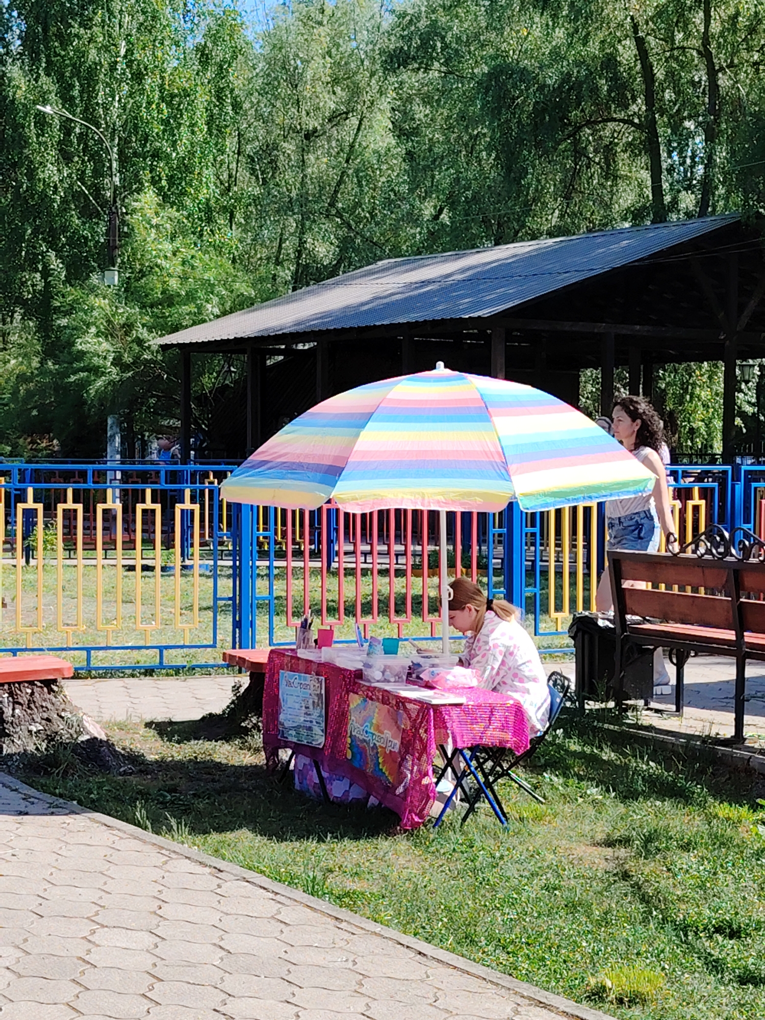 The image shows an outdoor scene in what appears to be a park or recreational area in Chaikovsky, Russia. A woman with curly dark hair and wearing a light-colored top and denim shorts is walking past a small table set up under a colorful striped umbrella. A younger person is seated at the table, possibly engaging in an activity such as painting or crafting, with various items on the table suggesting art supplies. The table is covered with a pink tablecloth. Behind the table is a colorful metal fence. A bench is visible to the right, and a covered structure is in the background. The area is bordered by paving stones and grass, with trees in the distance, indicating a park-like environment.
