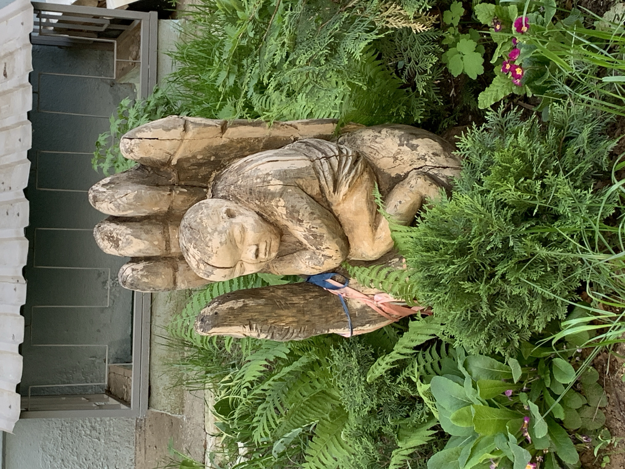 A sculpture of a sleeping figure nestled in a large hand, positioned amongst various green plants and flowers, stands near a set of stairs adjacent to a building. The sculpture appears to be made of wood. A blue and pink ribbon is tied around part of the hand. The setting is consistent with an outdoor area in a garden or park-like environment in Chaikovsky, Russia.
