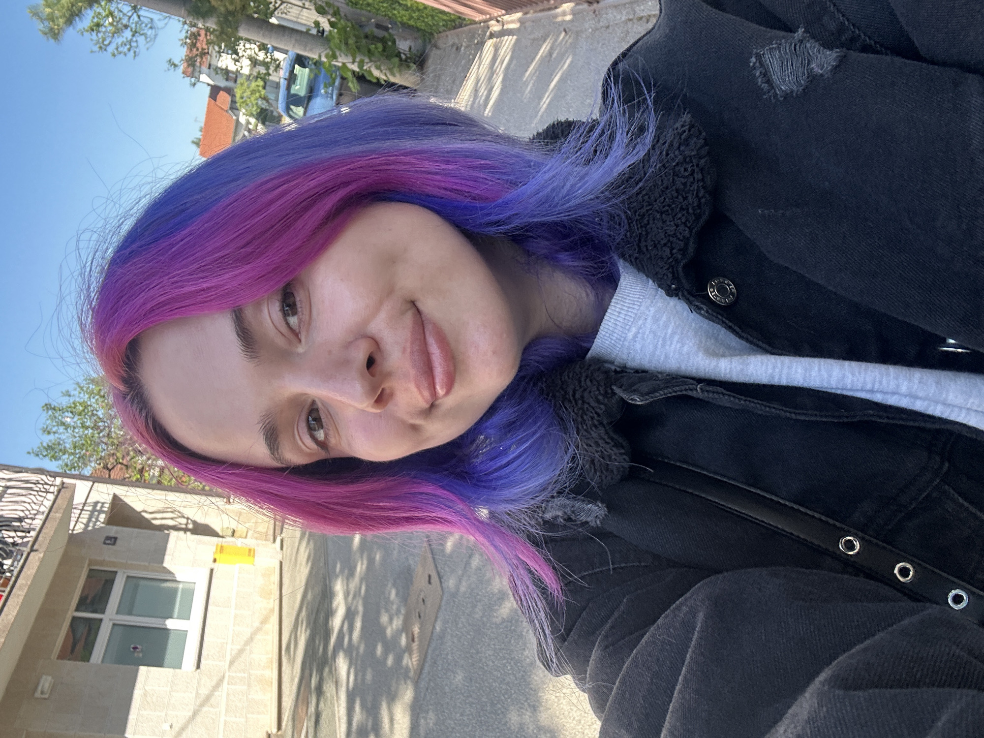A young woman with vibrantly colored hair, magenta on one side and purple-blue on the other, is featured in a selfie taken outdoors. Her long hair is styled and appears slightly windswept. She looks directly at the camera with a playful expression, a slight smile on her lips, and her tongue subtly extended. Her eyes are brown, and her eyebrows are dark.
She is wearing a light grey crew-neck shirt under a dark, possibly black, denim jacket. The jacket has distressed details, including rips, and a dark, fuzzy lining around the collar. A black strap with silver grommets is visible on her lower left, likely part of an accessory or bag.
The background suggests an urban or suburban environment in the City of Zagreb, Croatia, under a clear blue sky. Bright sunlight casts distinct shadows on the paved ground, indicating a sunny day. Buildings with varying facades, including a reddish-orange tiled roof and lighter-colored structures with windows, are visible. Green trees and a palm tree are scattered throughout the scene, and a light blue car is partially visible in the distance. No text is visible in the image.