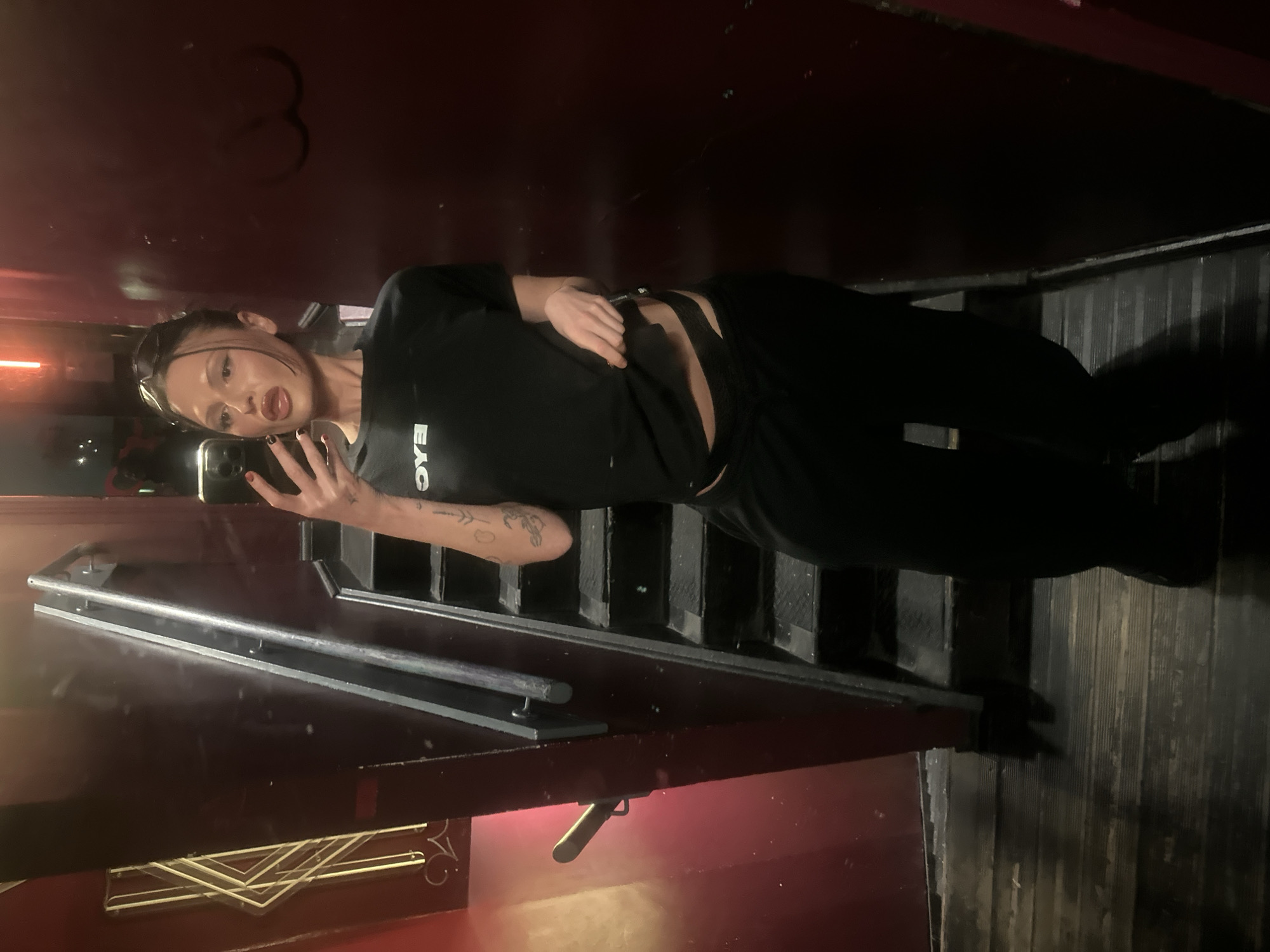 A female individual is depicted in a mirror selfie taken within the interior of The Fitzgerald bar, 11 Stevenson Square, Manchester M1 1DB, United Kingdom. She stands on dark wooden floorboards, adjacent to a dark, textured staircase with a metallic railing ascending on the right. The surrounding walls are dark red. On the left wall, a geometric yellow neon light fixture, consistent with Art Deco design, is visible above a brass-colored handle.
The individual, with dark hair pulled back and sunglasses resting on her head, is wearing a black short-sleeved t-shirt with white text (partially visible, appearing as 'OYE' or 'OYA') on the chest. She wears black loose-fitting trousers and black high-cut underwear, which is exposed as she lifts her t-shirt hem with her right hand. Her left hand holds a black smartphone, capturing her reflection. She has dark fingernail polish and two distinct tattoos on her left forearm. An orange-red light source is visible near the top center of the frame, contributing to the dim ambient lighting.