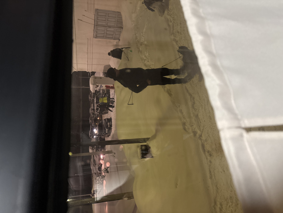 A person stands on snow-covered ground, reflected in a window. In the background, a building with closed doors is visible along with parked vehicles. Other people are present in the scene, also on the snow. The reflection obscures the view, creating a layered visual effect. The lighting suggests it is nighttime or a dimly lit environment. A white curtain is partially visible in the right side.