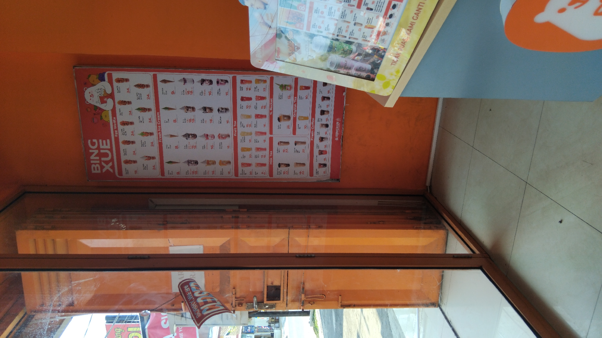 The image depicts the exterior of a BINGXUE shop in Batang, Indonesia. A bright orange wall forms the backdrop for a menu board displaying various drinks and ice cream items. The menu is categorized into "Egg Waffle," "Fresh Ice Cream," "Fruit Tea," "Milk Tea," "1L Bucket," "Original Tea," and "Fresh Coffee." Prices are listed in Indonesian Rupiah (Rp).
To the left, a glass door and orange-colored wall of the shop are visible. A red and white "SORRY, WE ARE CLOSED" sign hangs on the door. The floor is tiled with light-colored square tiles. In the foreground, a display stand with a menu is positioned to the right, showing more BINGXUE products and promotional text in Indonesian, "TILAK PUAS, KAMI GANTI."
The overall scene suggests a commercial establishment, likely a fast-food or dessert shop, on a day when it is closed for business. The environment appears to be outdoors, with a hint of a street visible through the glass door.