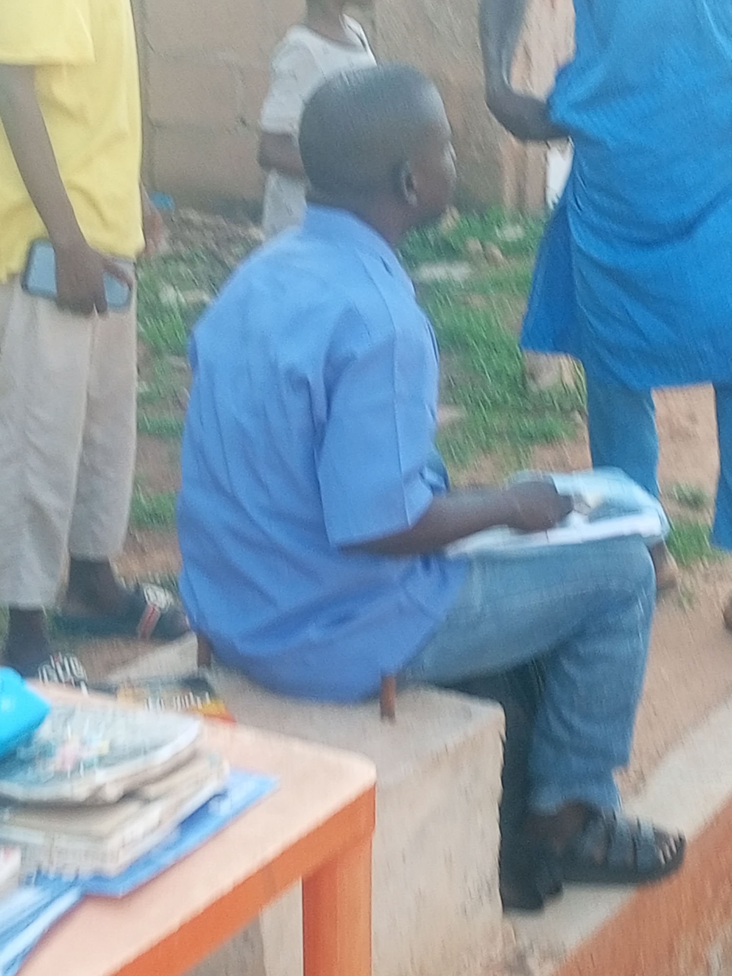 The image depicts an outdoor scene, likely in Gombe, Nigeria, characterized by a slightly blurred, grainy quality. In the foreground, a person is seated, wearing a blue collared shirt and blue denim jeans. Their right hand is holding an open book or document. To the left, another person is visible, wearing a bright yellow t-shirt and light-colored pants, holding a smartphone in their left hand. A portion of a person in a blue garment is visible in the upper right. The environment appears to be unpaved, with patches of grass and dirt visible. A simple orange table with stacked books and what appears to be a blue object is positioned in the lower left corner. The overall impression is of a casual gathering or a lesson taking place outdoors.