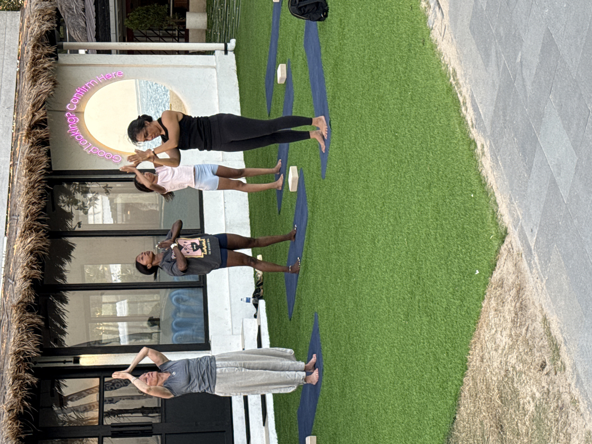 An outdoor yoga session is taking place on a patch of artificial turf in front of a building with a thatched roof and large windows. Four individuals, three women and one girl, are standing on yoga mats, facing forward with their hands in prayer position at chest level. A neon sign above them reads "Good Looking? Confirm Here" in a circular arc. The building's white facade is visible, along with a large circular window revealing a water view. In the background, trees and more of the building's structure are present. The ground is a mix of green turf and sand, with a paved walkway visible in the foreground. This scene is located in Denpasar, Indonesia.