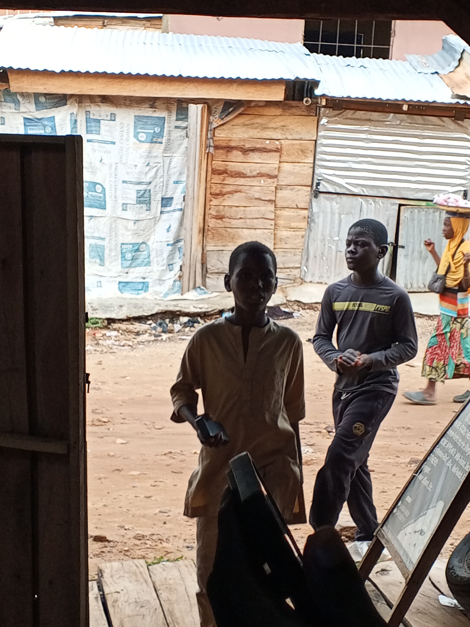 Outside a wooden building in Lokoja, Nigeria, two boys are walking down a dirt path. The boy in front is wearing a tan tunic and holding a small object in his right hand. Behind him is a boy wearing a long-sleeved gray shirt and dark pants with a small design on the left thigh. A woman wearing a yellow hijab and a colorful skirt walks in the background carrying a tray on her head. Beside the path, to the right, is a display board with writing on it. The buildings are simple structures with wood or corrugated metal.